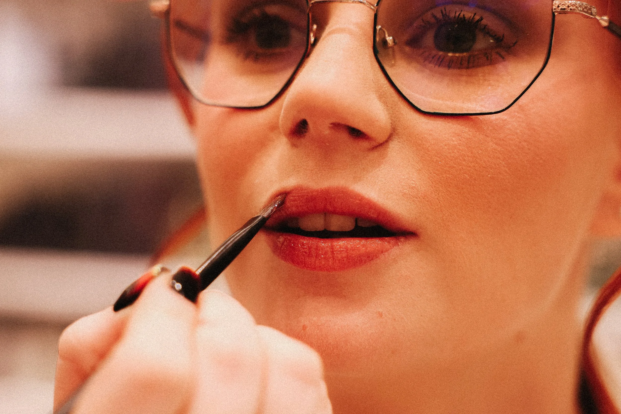 Une femme portant des lunettes applique du rouge à lèvres avec un pinceau, concentrée sur son maquillage.