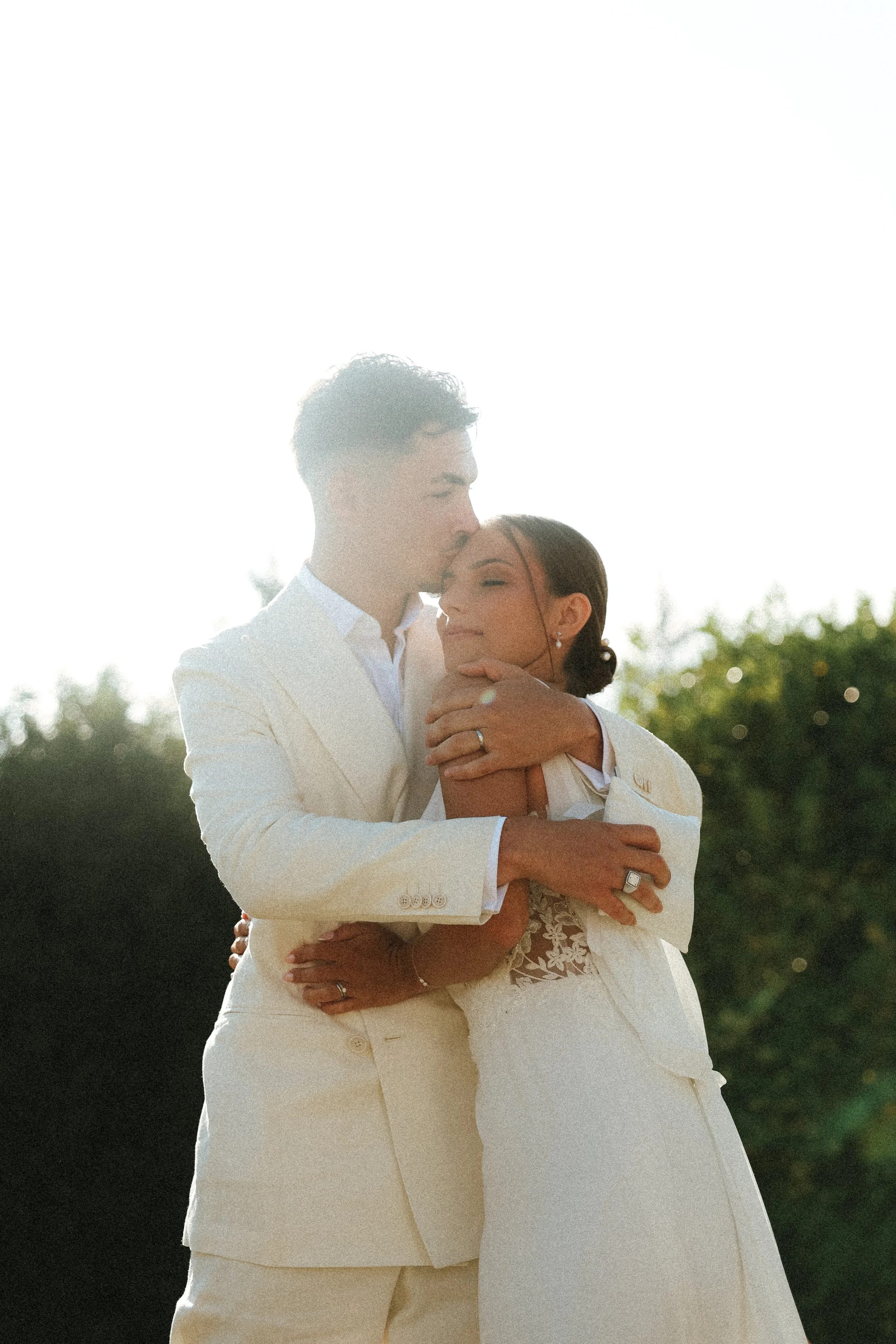 Un couple en tenue de mariage s'embrasse tendrement à l'extérieur avec un arrière-plan de verdure, sous la lumière du soleil.
