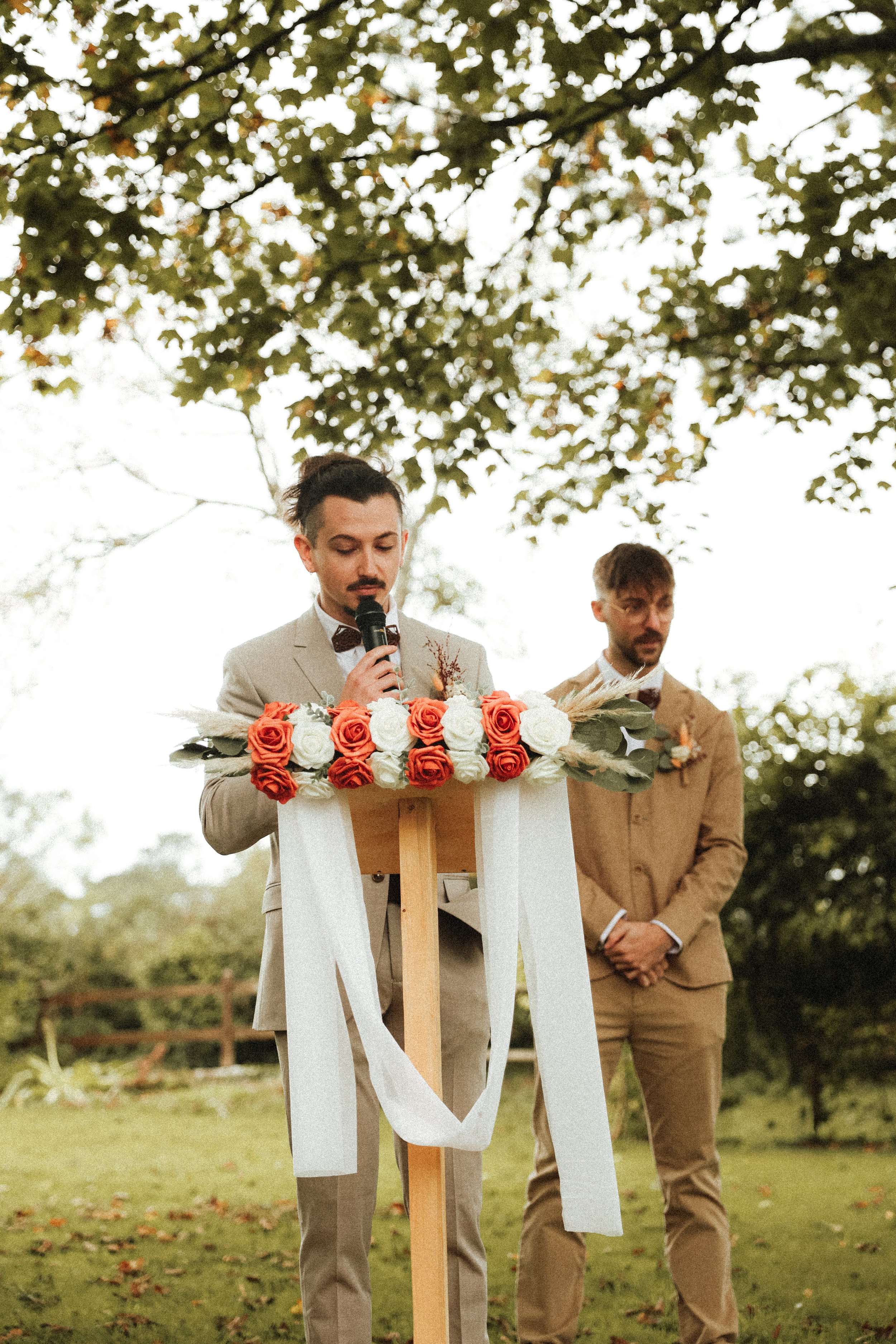 Deux hommes en costume en plein air, l'un parlant au microphone lors d'une cérémonie, sous un arbre aux feuilles d'automne.