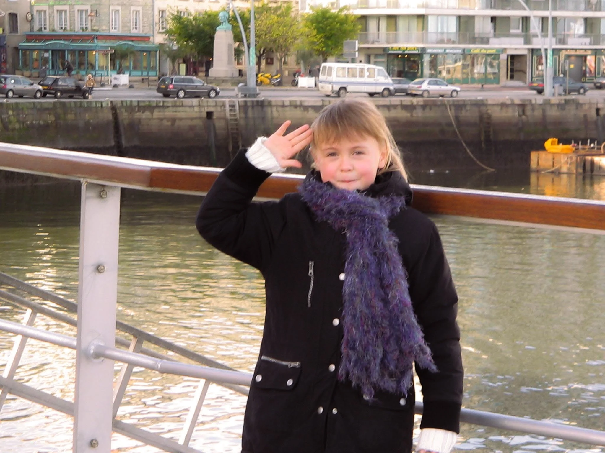 Jeune fille saluant tout en se tenant près d'un quai le long de l'eau, avec des voitures et des bâtiments en arrière-plan.