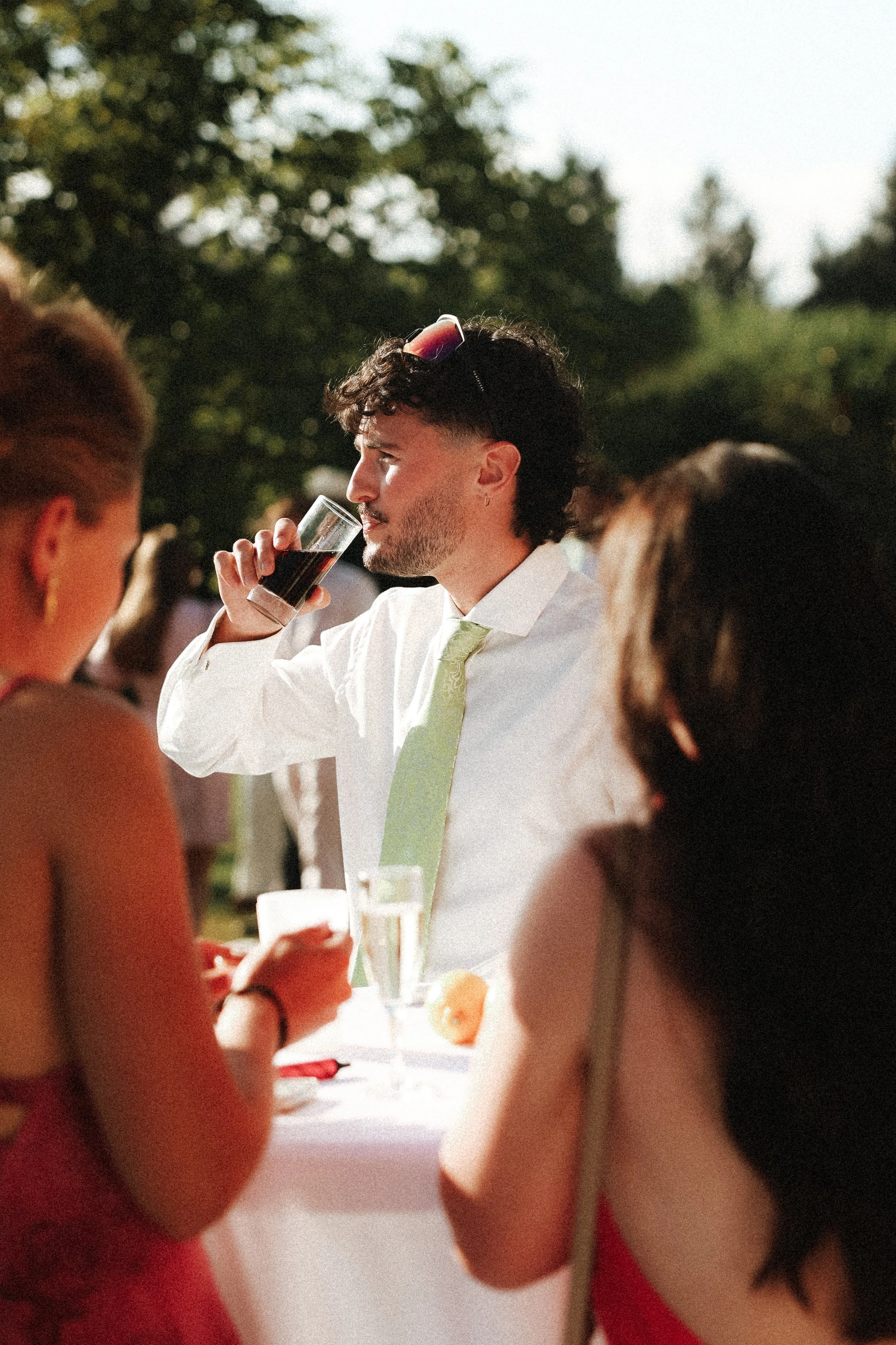 Un homme avec des cheveux bouclés, portant une chemise blanche et une cravate verte, boit un verre de boisson sombre lors d'un événement en plein air, entouré de personnes, avec des arbres en arrière-plan.