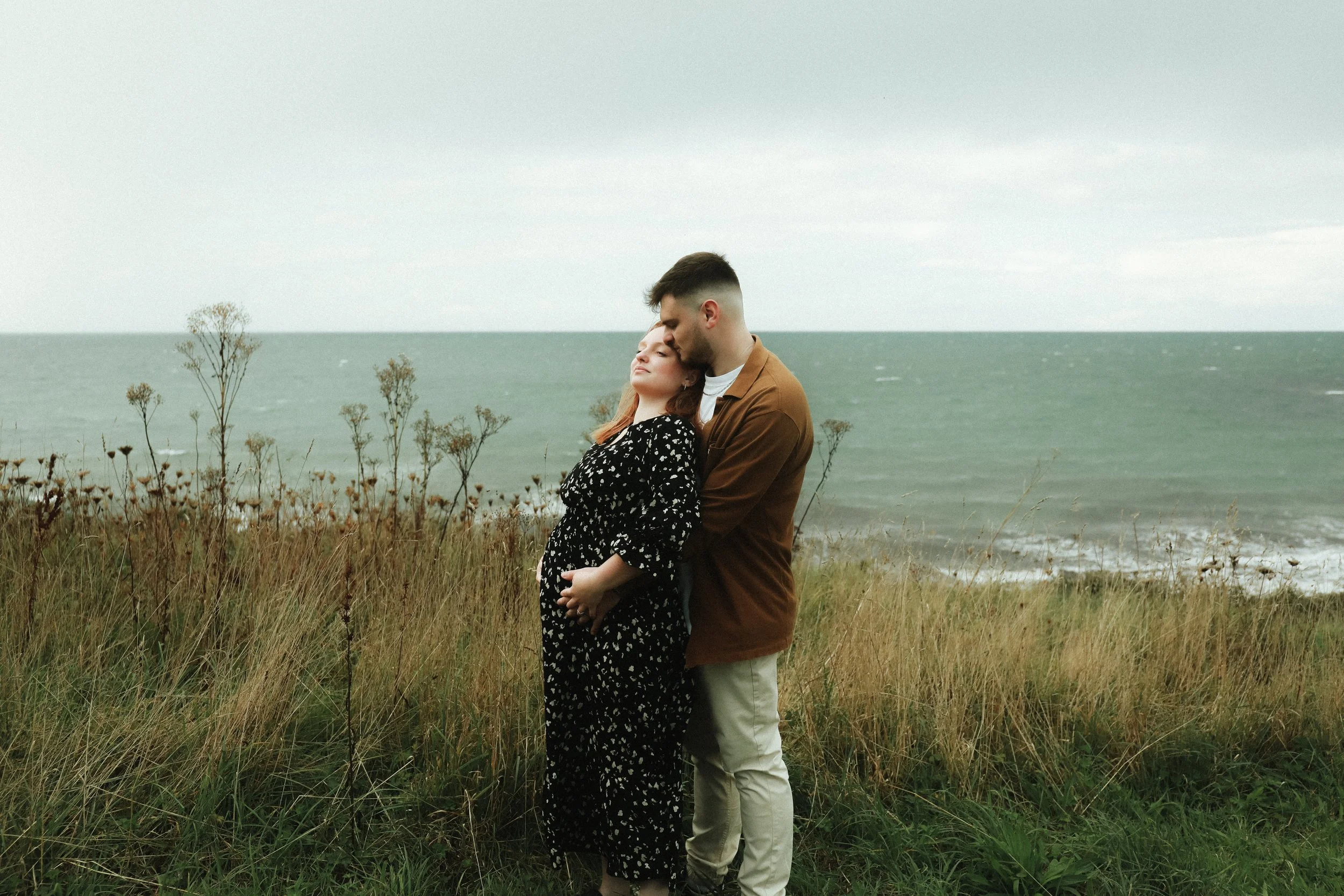 Un couple debout dans un champ près de la mer, la femme enceinte embrassant la tête de l'homme.
