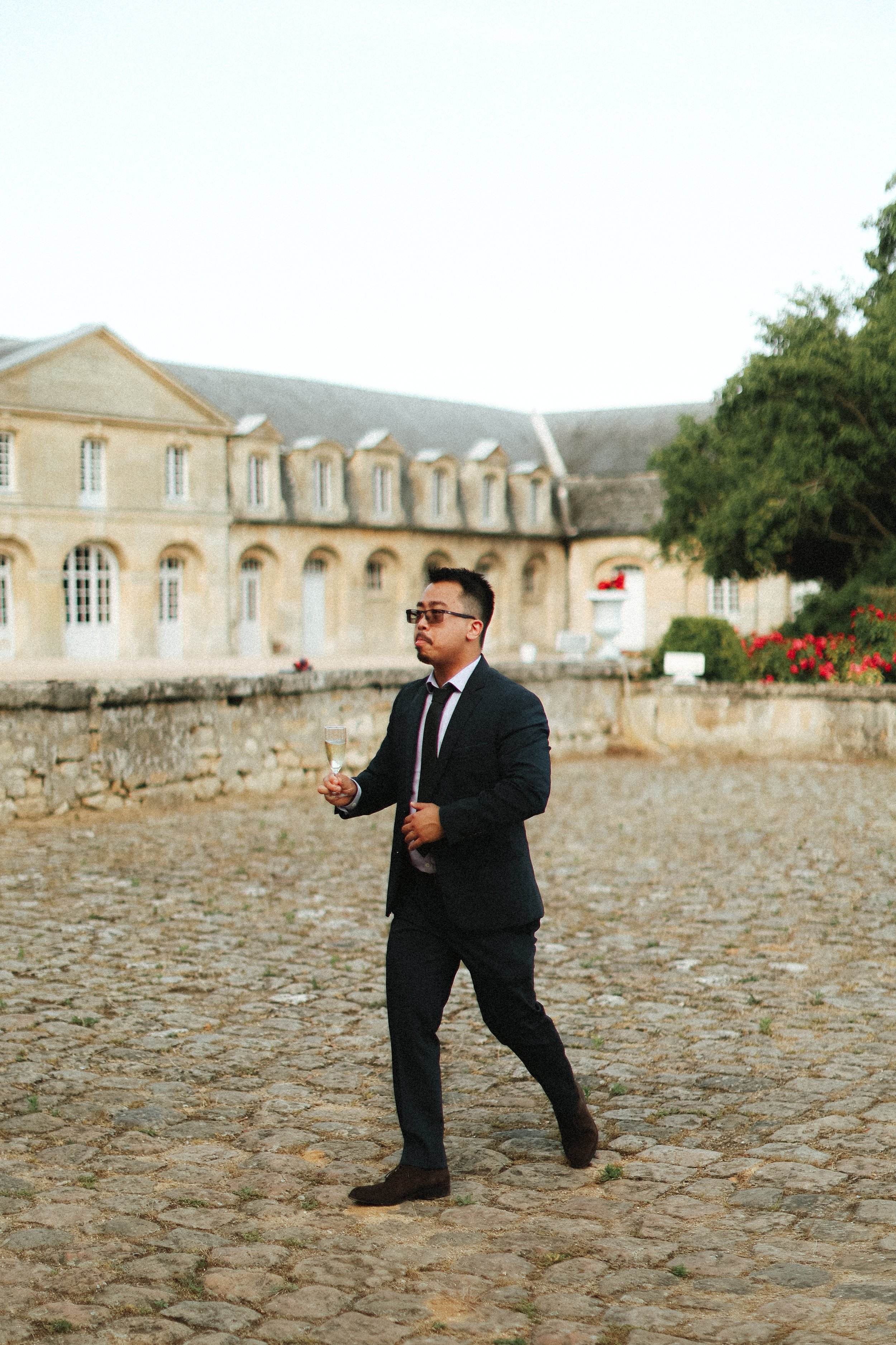 Un homme d'affaires en costume noir marche sur une cour pavée avec un verre de champagne à la main, devant un bâtiment historique en pierre.