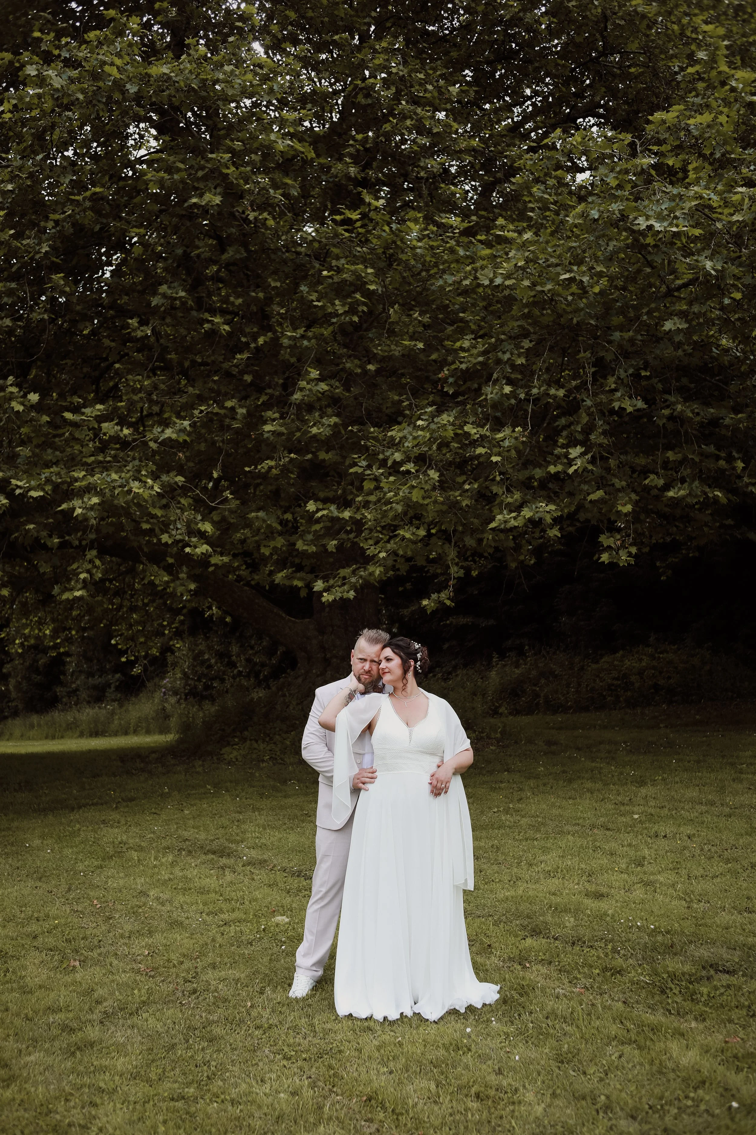 Un couple en mariage posant devant un arbre dans un parc, l'homme en costume blanc et la femme en robe blanche, photographe Normandie