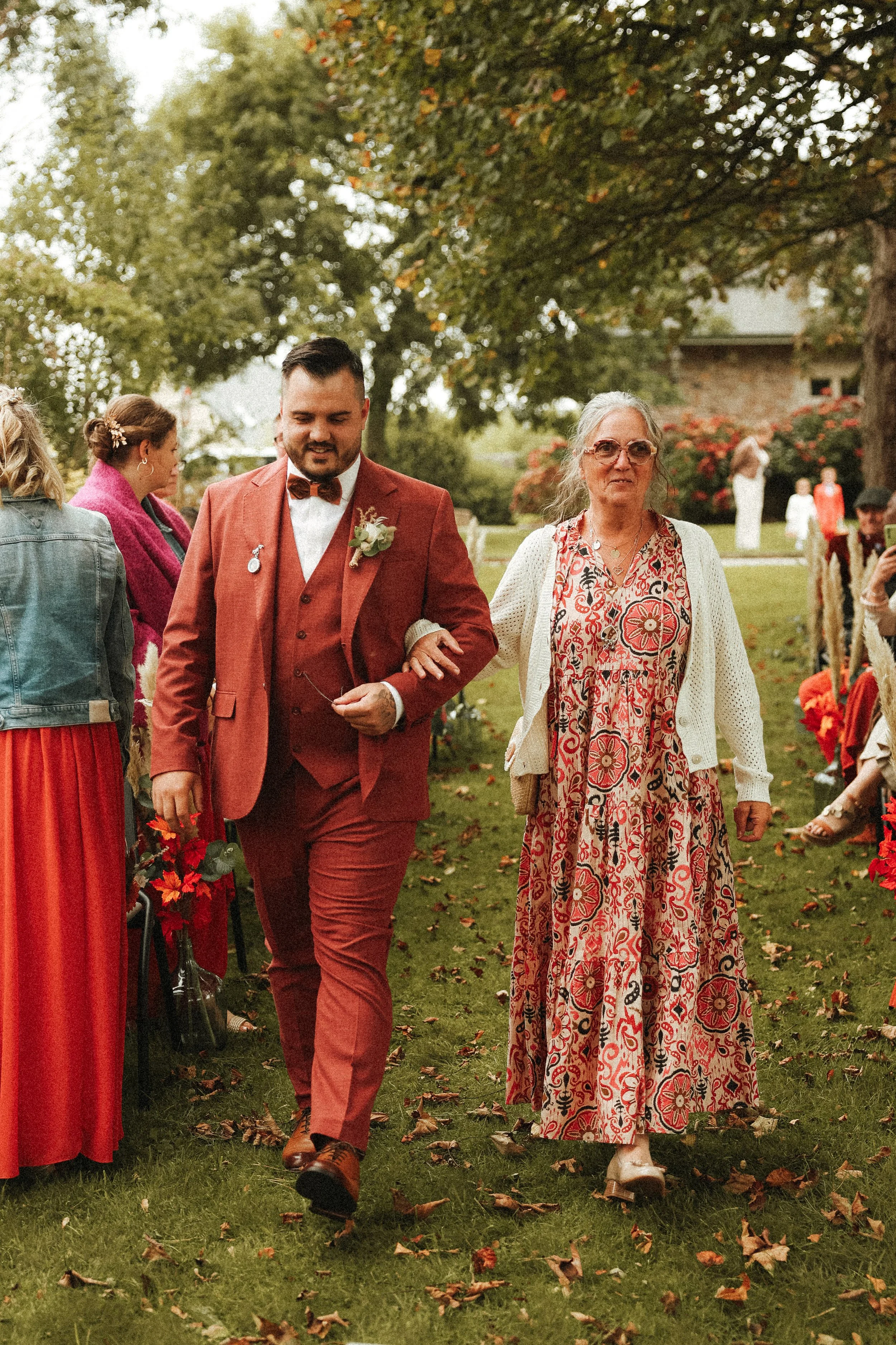 Un homme en costume rouge avec une official piqûre d'honneur et une femme en robe longue à motifs floraux marchent ensembles lors d'un mariage en plein air, entourés d'invités et de nature automnale.