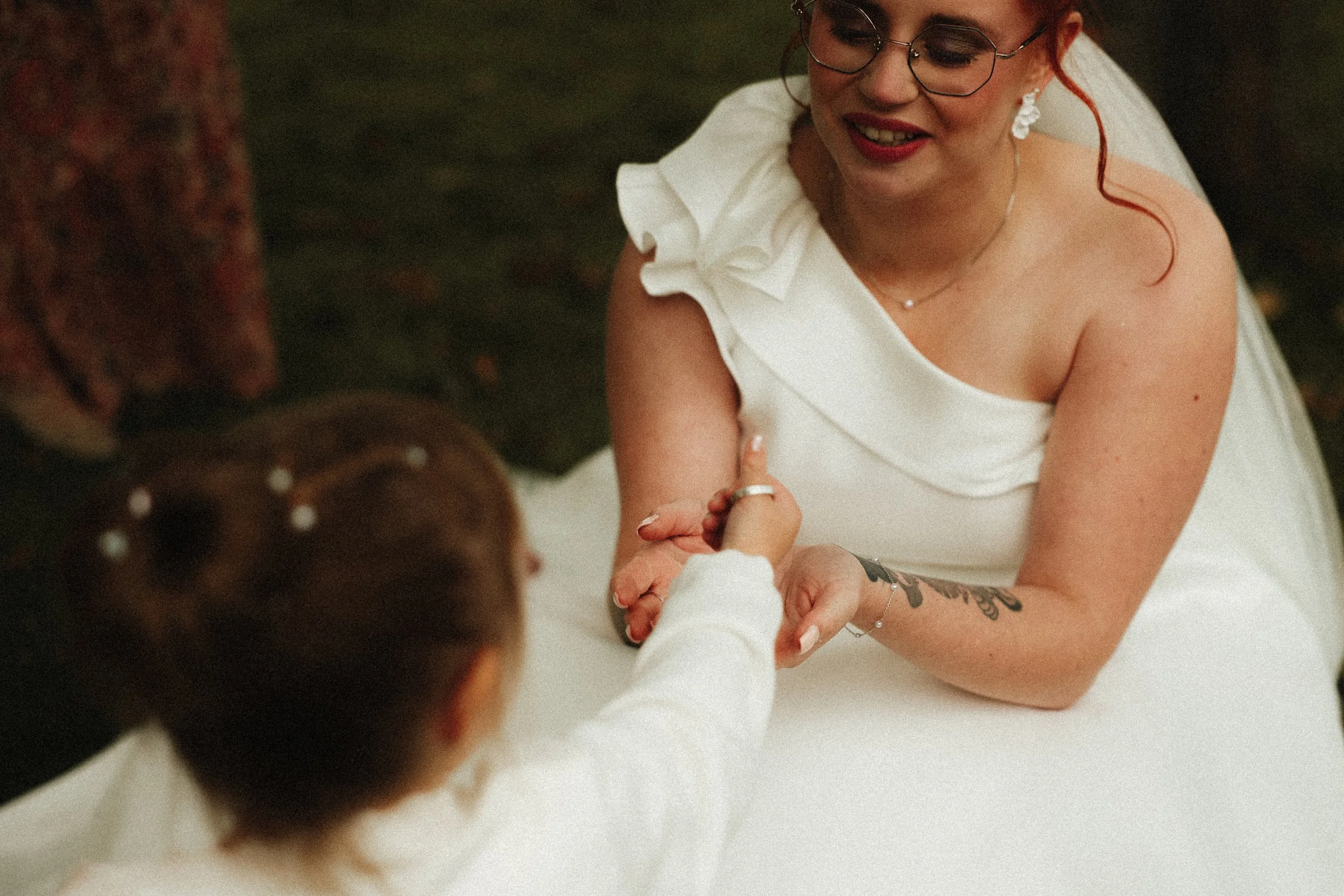 Une femme en robe blanche, portant des lunettes et des bijoux, sourit en tenant la main d'une enfant avec qui elle partage un moment d'émotion. L'enfant a les cheveux à petits points blancs et porte une veste blanche.