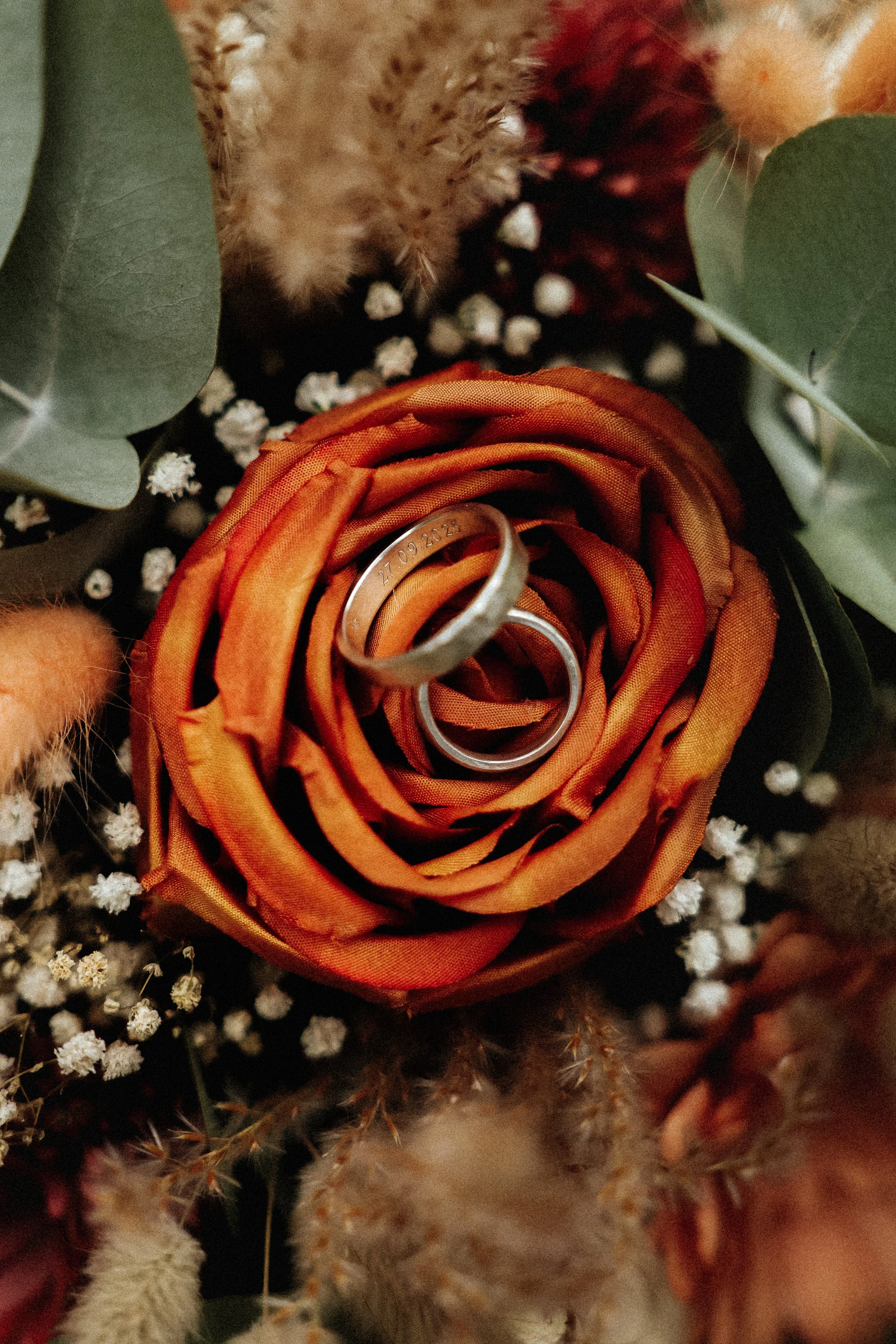 Un bouquet de fleurs avec deux alliances en métal argenté placées au centre d'une rose orange avec des feuilles vertes et des petites fleurs blanches autour.