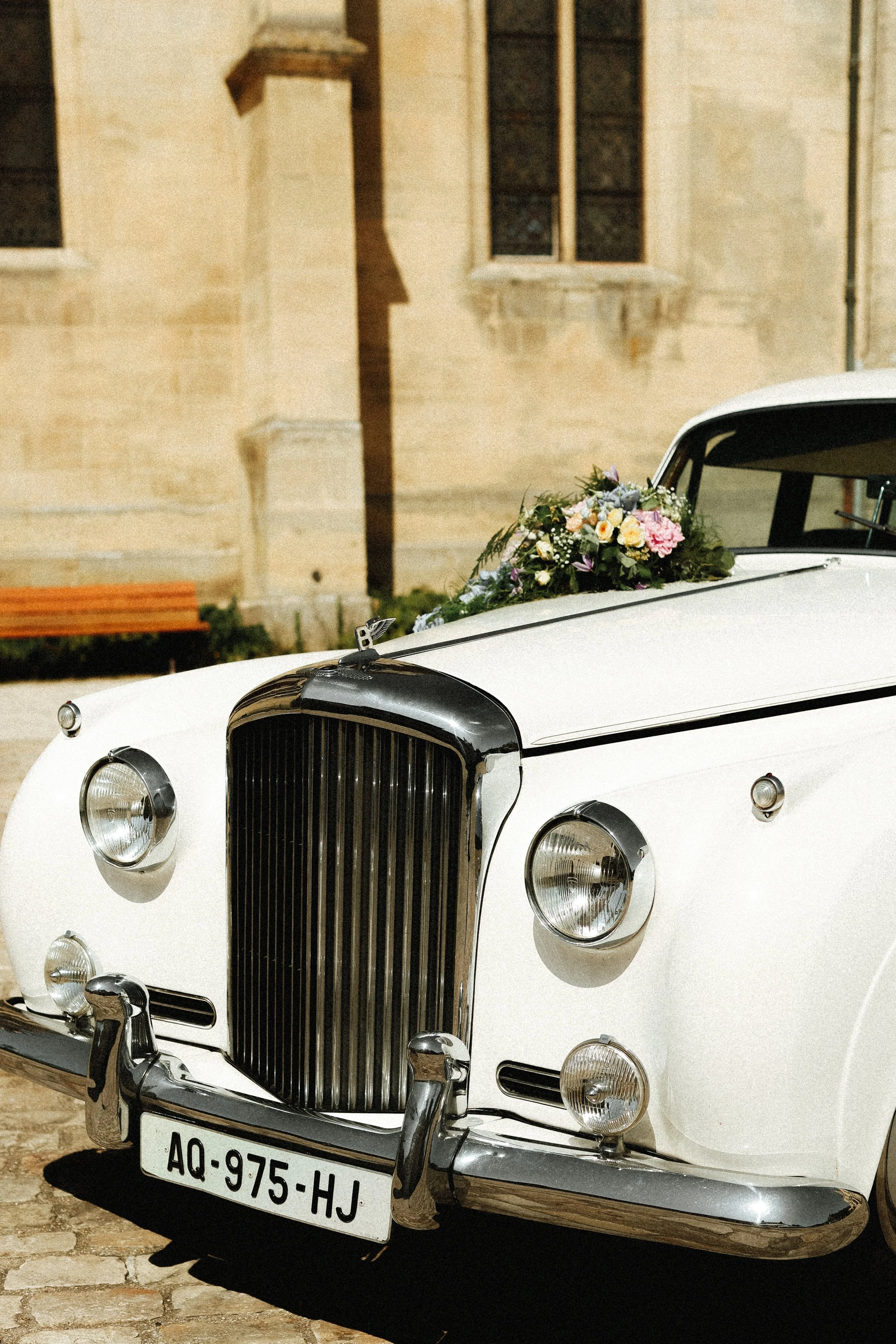 Voiture ancienne blanche avec une décoration florale sur le capot, stationnée devant une façade en pierre d'un bâtiment historique.