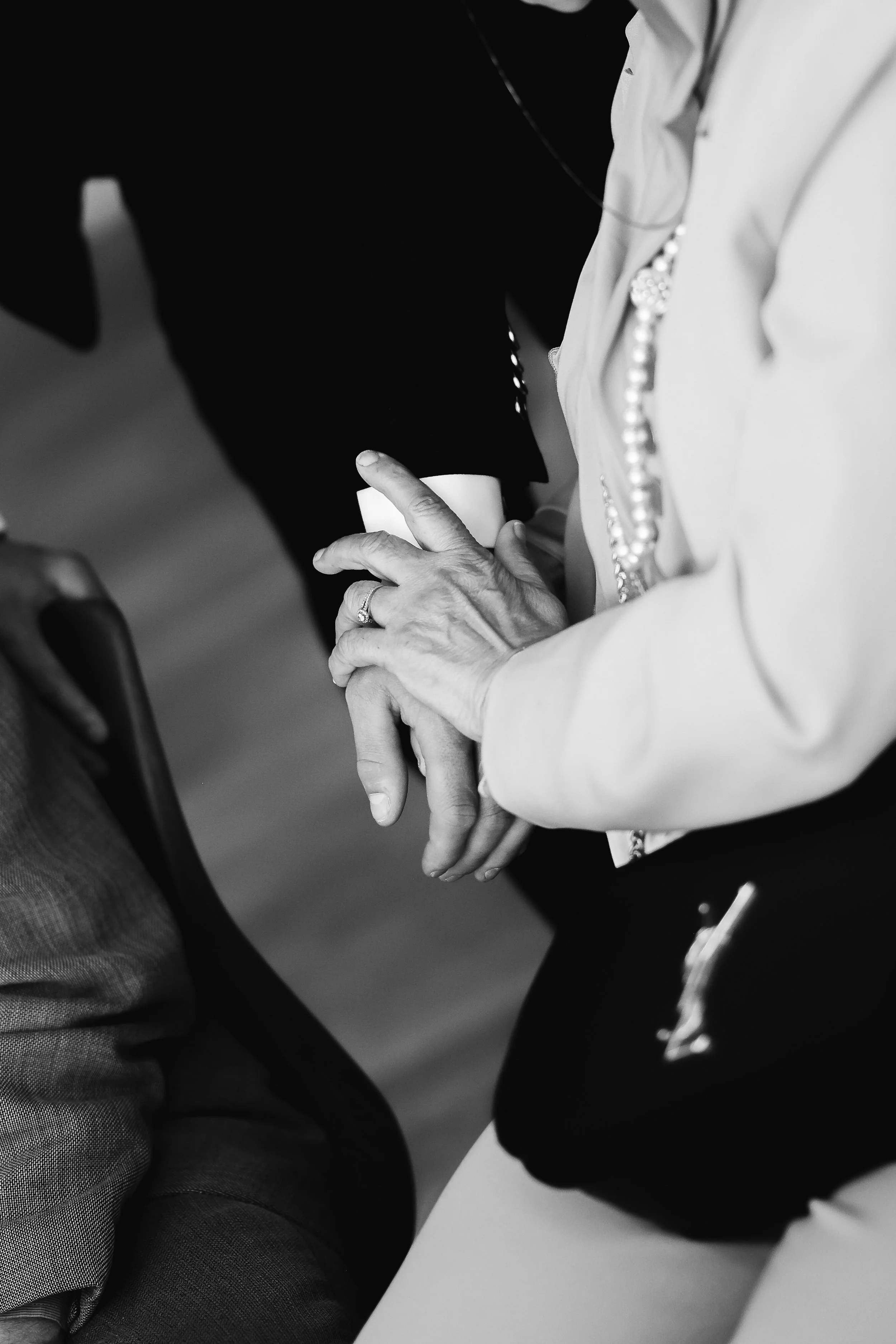 Une personne âgée avec des mains visibles, portant une chemise blanche avec des perles, tenant une tasse. La scène semble être dans un environnement intérieur avec d'autres personnes en arrière-plan.