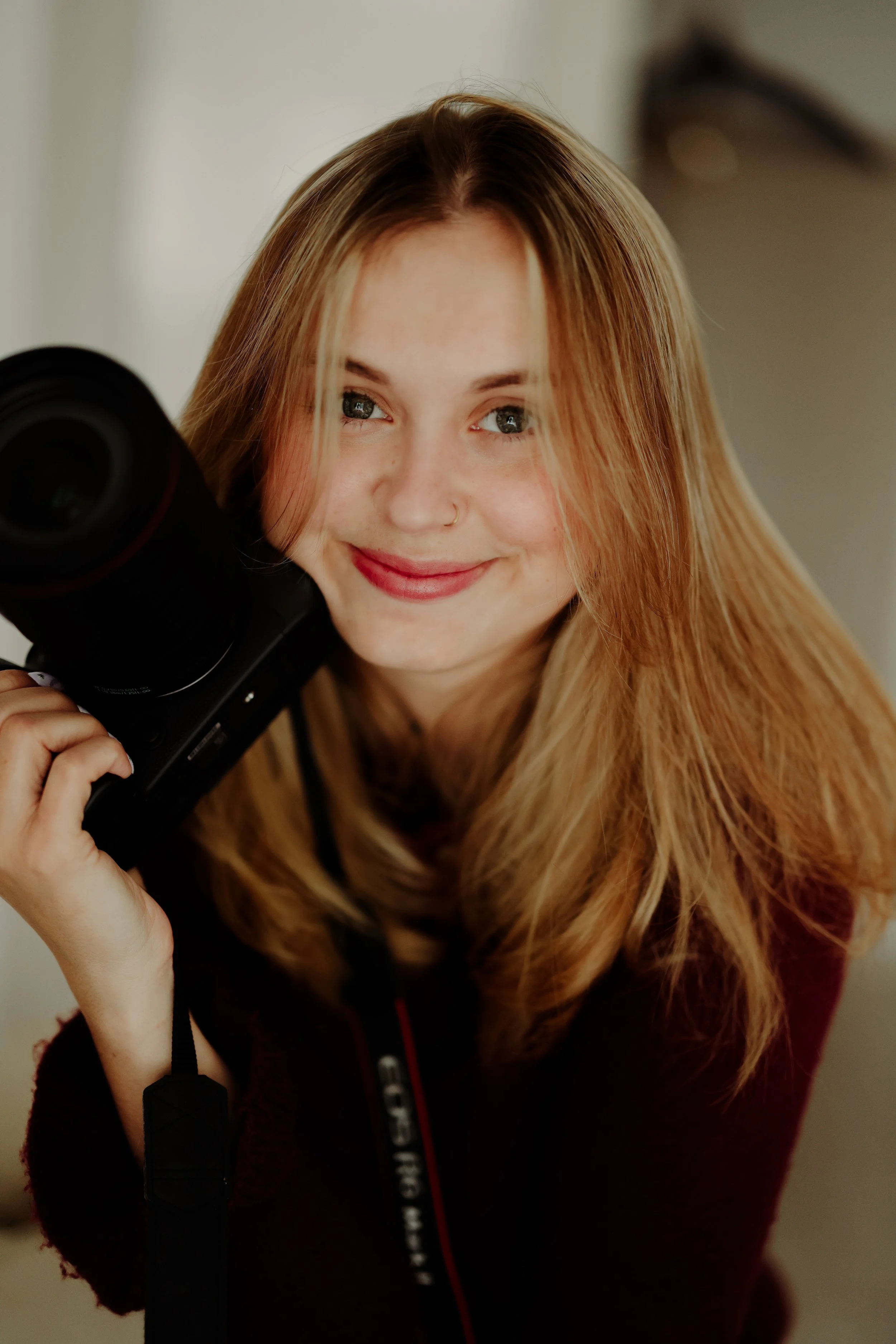 Jeune femme aux cheveux roux, souriante, tenant un appareil photo dans sa main, portant un sweat noir avec une fermeture éclair rouge.