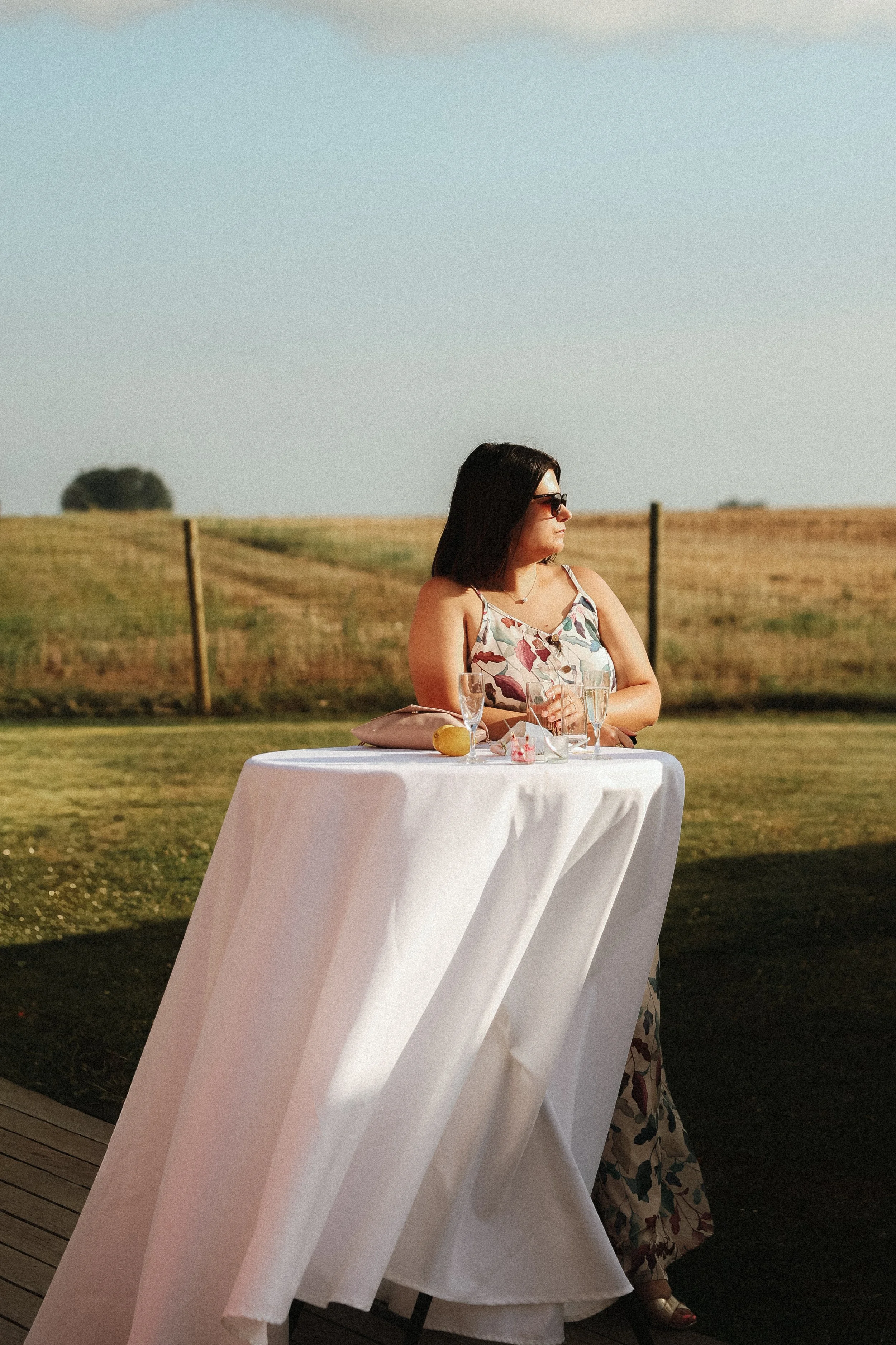 Une femme portant une robe florale noire, assise à une table couverte d'une nappe blanche à l'extérieur, avec un champ en arrière-plan, buvant un verre de champagne.