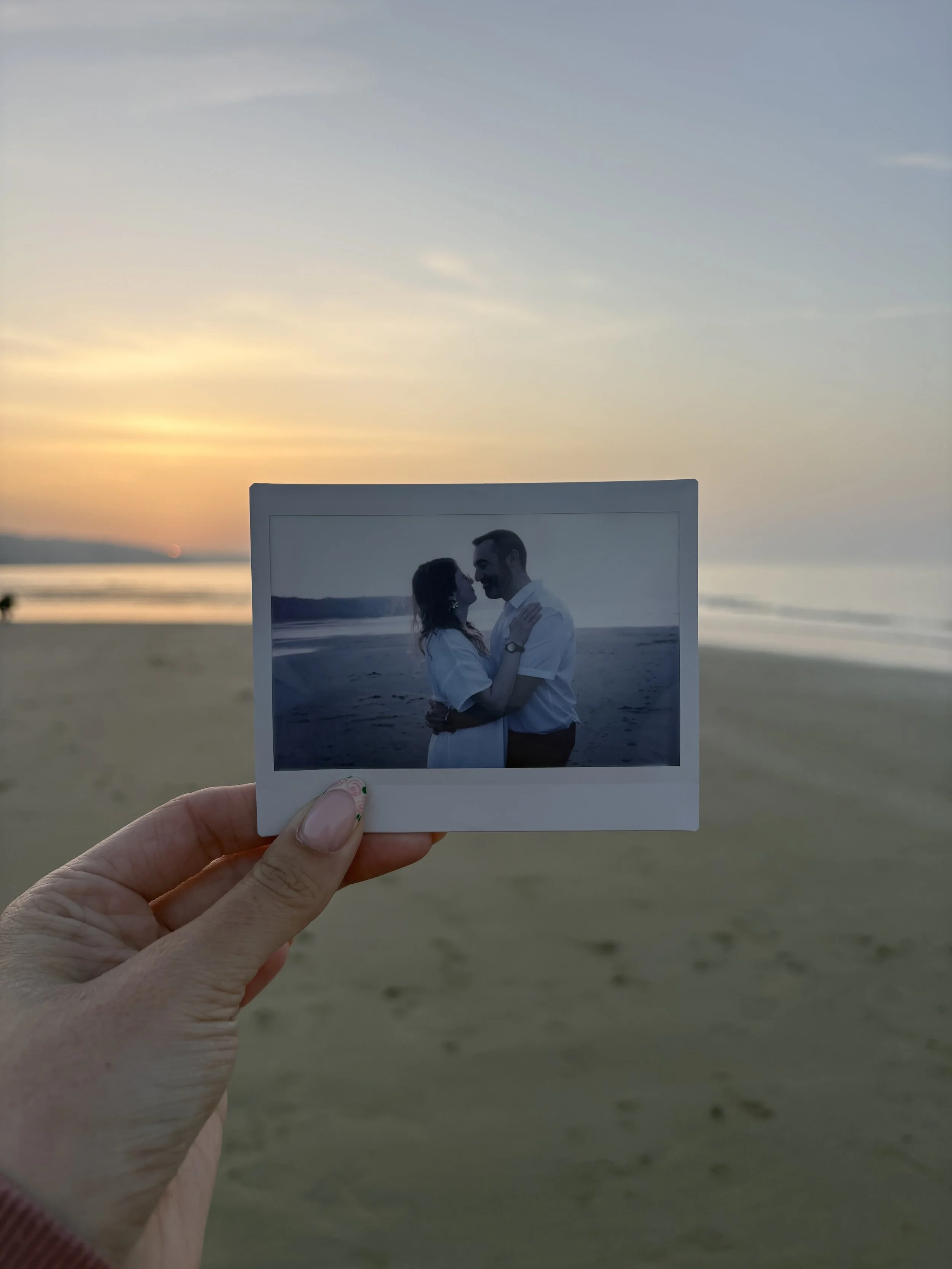 Une personne tient une photo argentique d'un couple s'embrassant sur la plage au coucher du soleil.