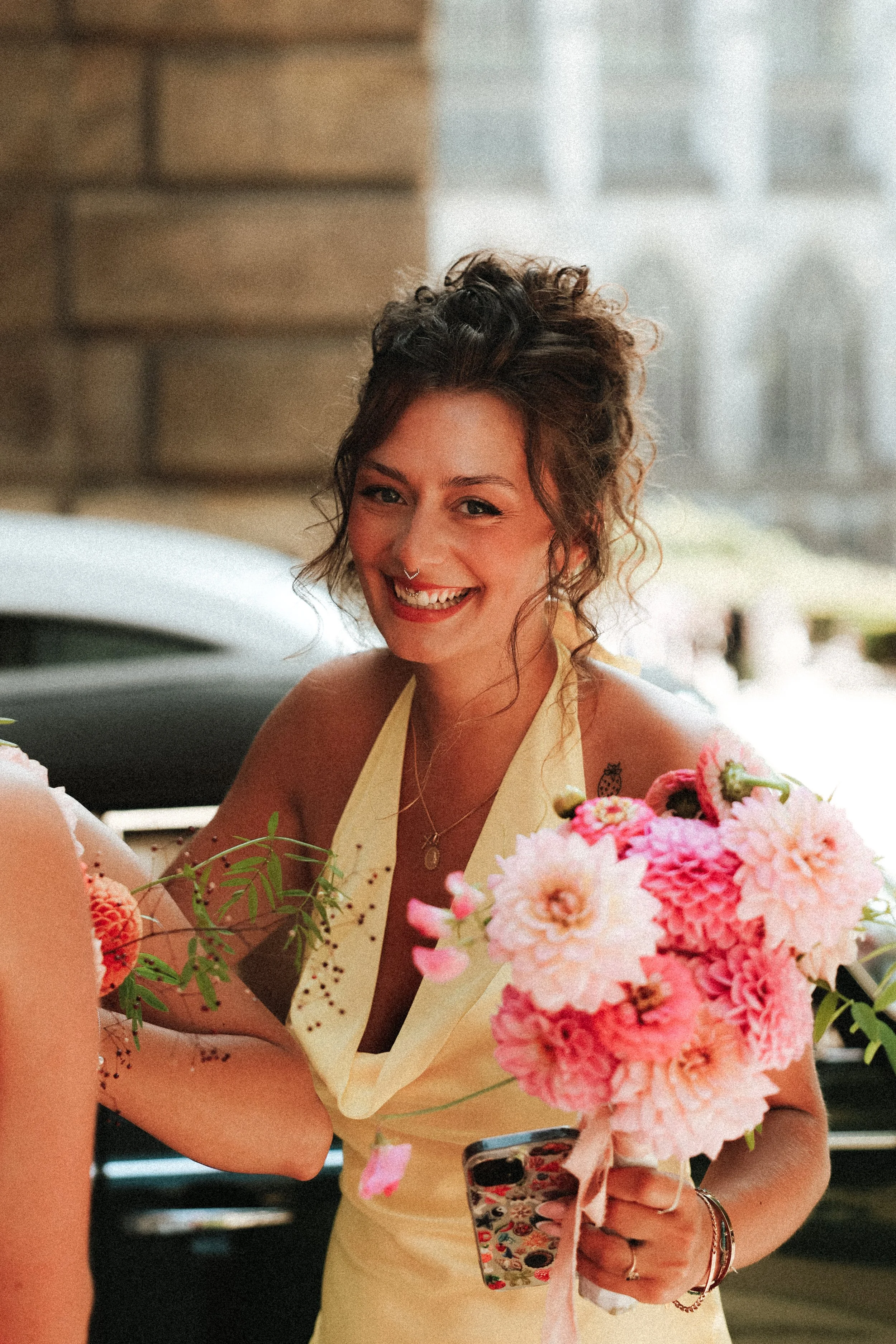 Une femme souriante avec des cheveux bouclés, portant une robe jaune, tenant un bouquet de fleurs roses, décorant avec un tatouage sur l'épaule, en extérieur avec un bâtiment en arrière-plan.