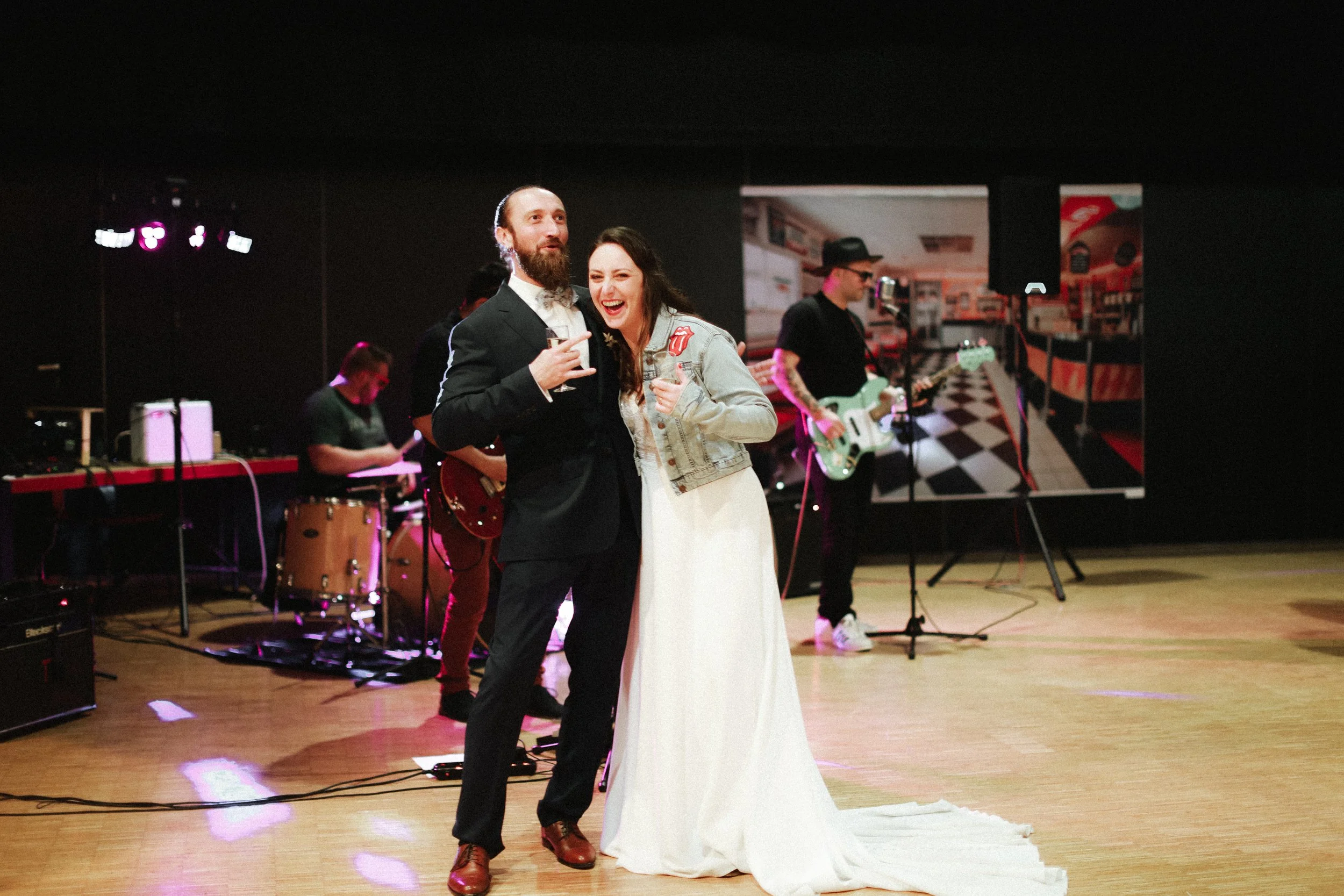 Deux personnes souriantes, un homme en costume et une femme en robe blanche, posent lors d'une célébration avec un groupe de musiciens en arrière-plan