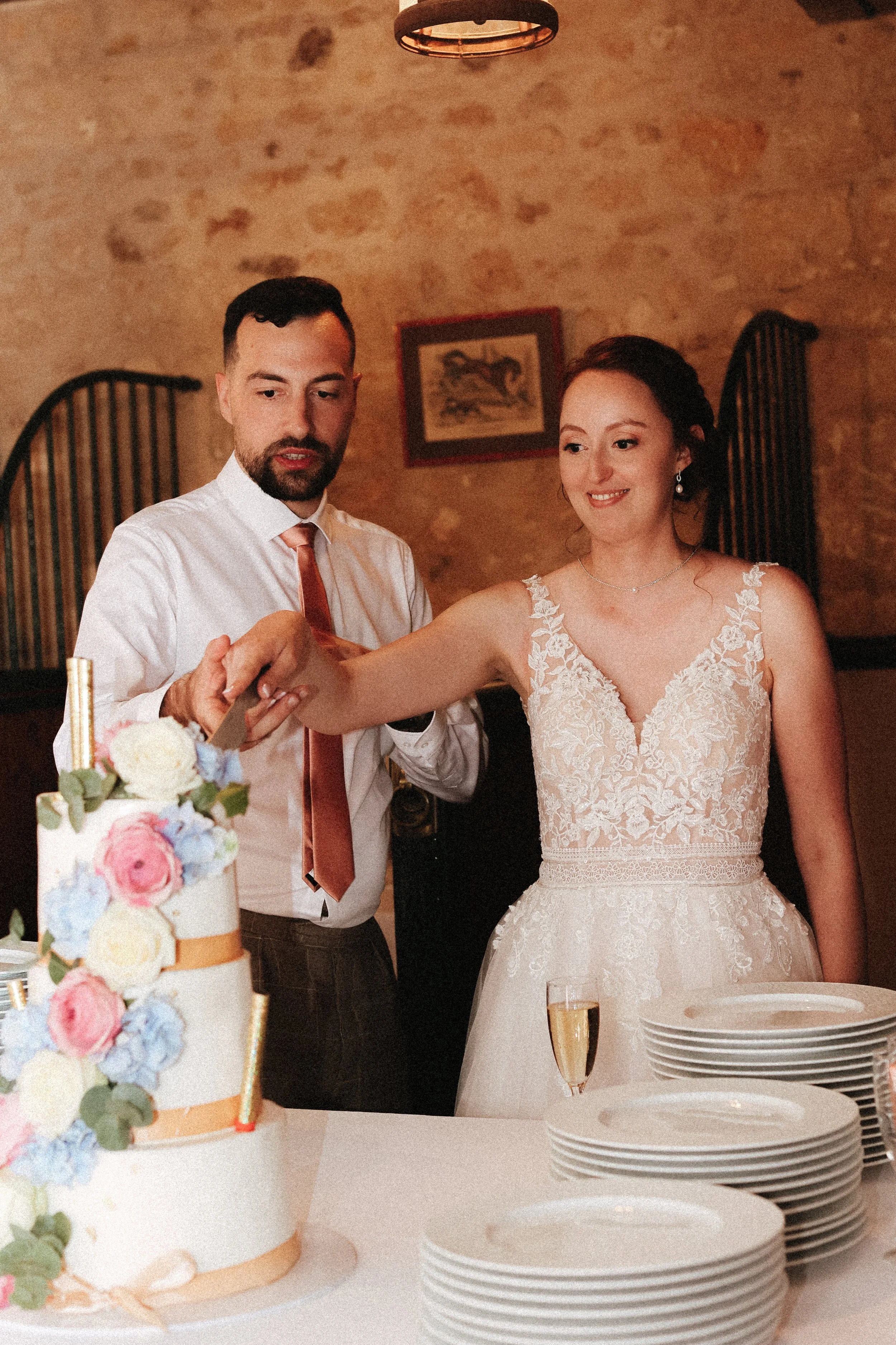 Un couple lors de leur mariage coupe un gâteau de mariage décoré de fleurs, avec des assiettes et des verres de champagne sur la table.