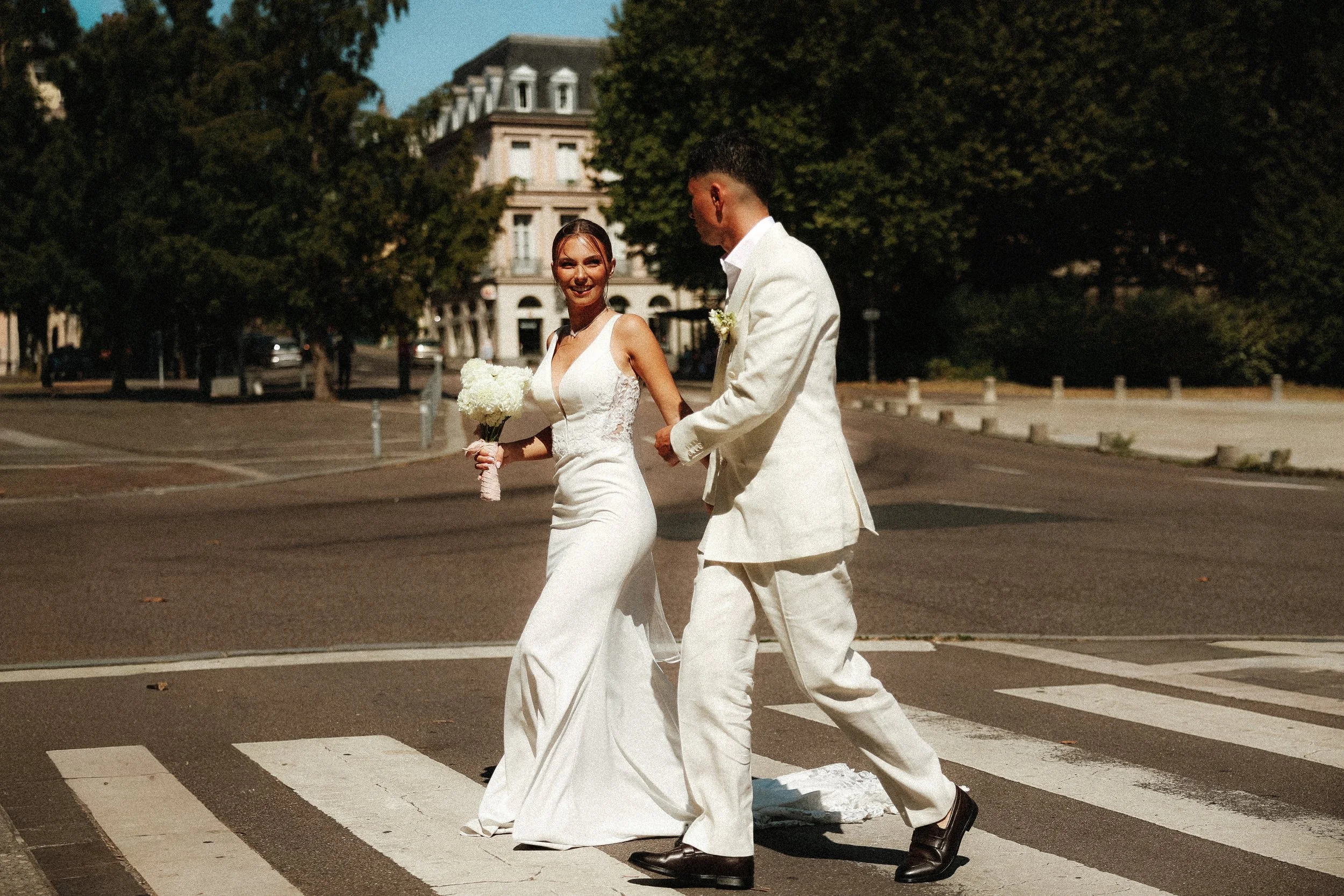 Un couple de mariés marchant sur un passage piéton dans une rue urbaine, la femme portant une robe de mariée blanche et tenant un bouquet de fleurs, l'homme en costume blanc, avec des arbres et des bâtiments résidentiels en arrière-plan.