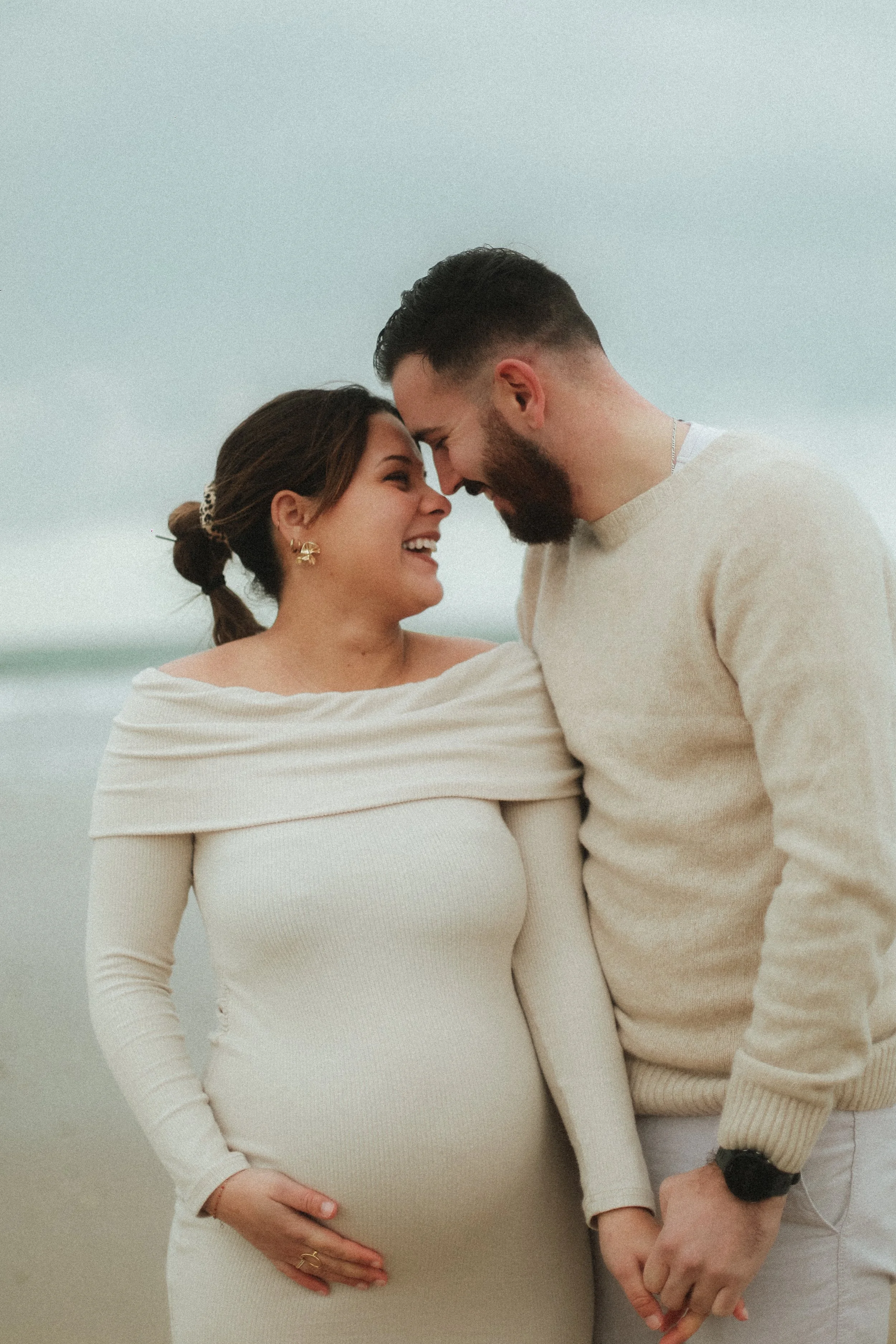 Un couple souriant, où la femme est enceinte, se tenant la main sur la plage, avec des vêtements beiges et des cheveux bruns, échangeant un regard heureux par temps nuageux.