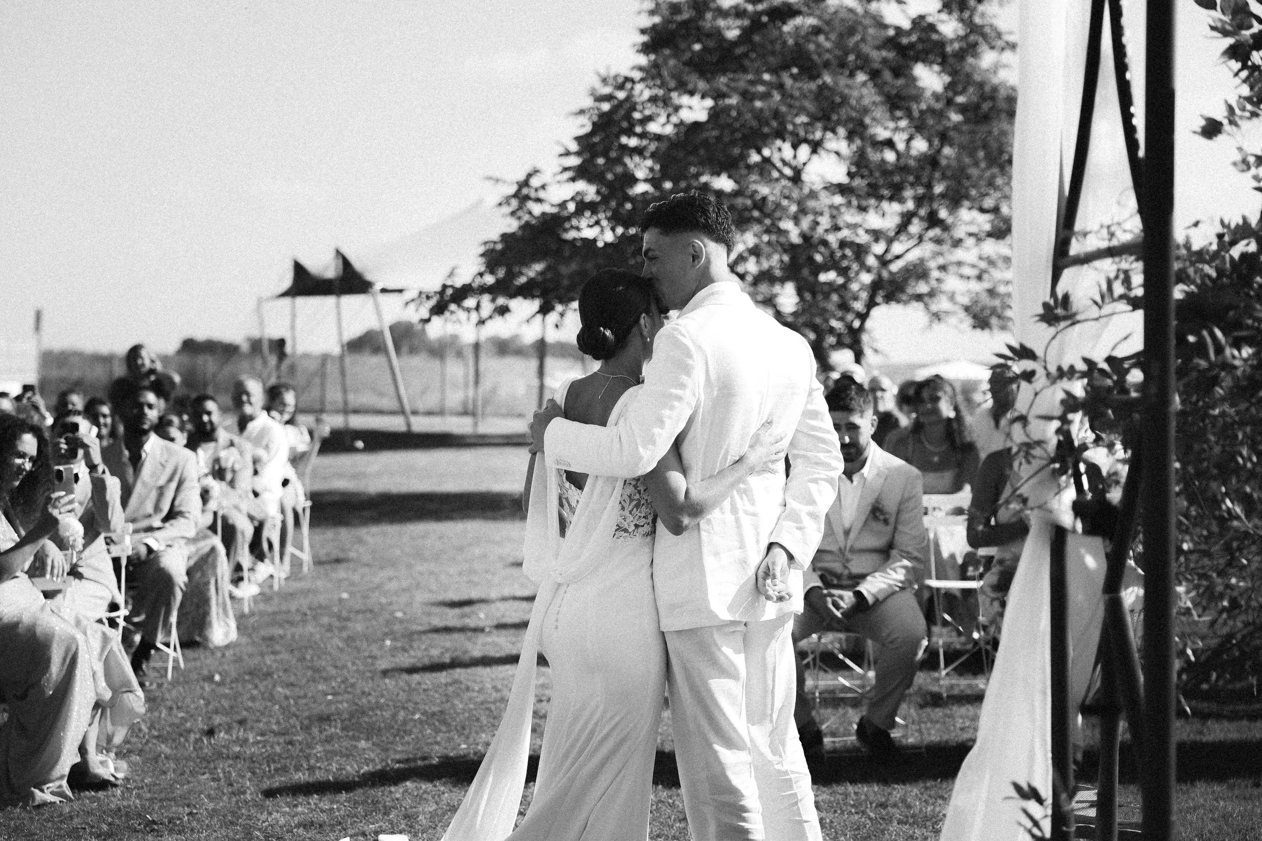 Un couple en mariage partage une danse lors de leur cérémonie en plein air, entouré d'invités assis et d'arbres, en noir et blanc.