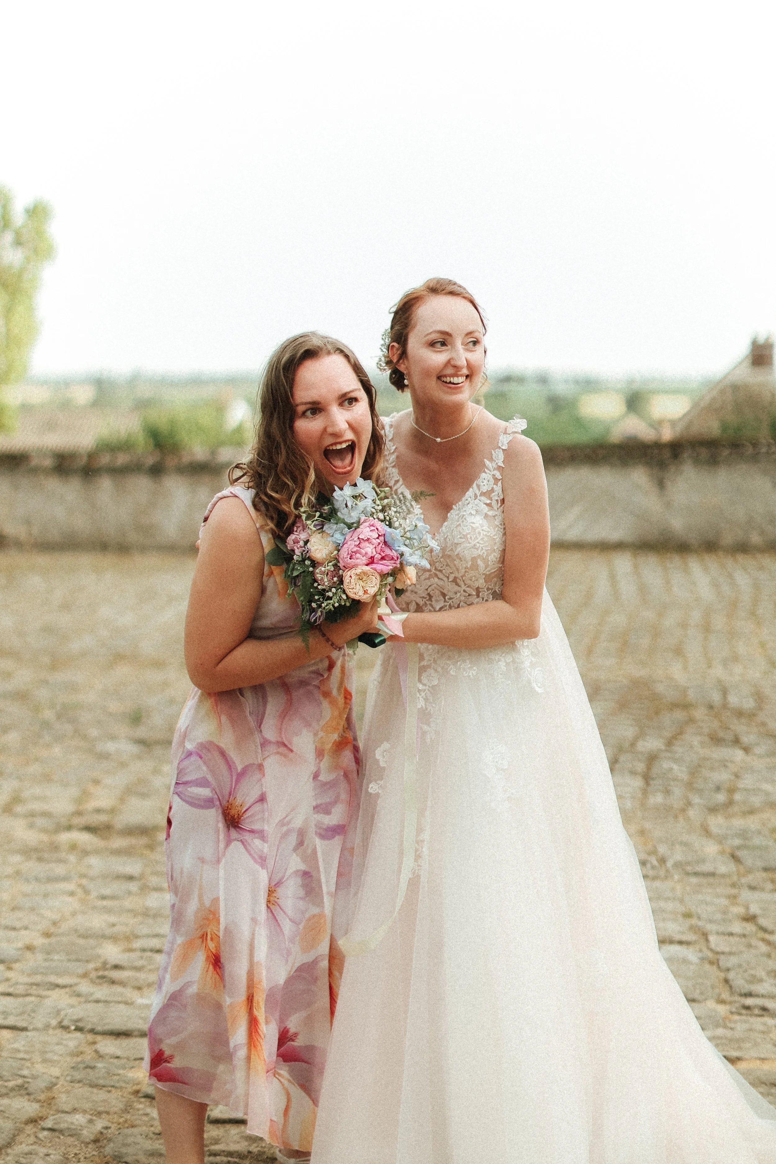 Deux femmes souriantes, dont une en robe de mariée blanche, tenant un bouquet de fleurs, lors d'un mariage en plein air.