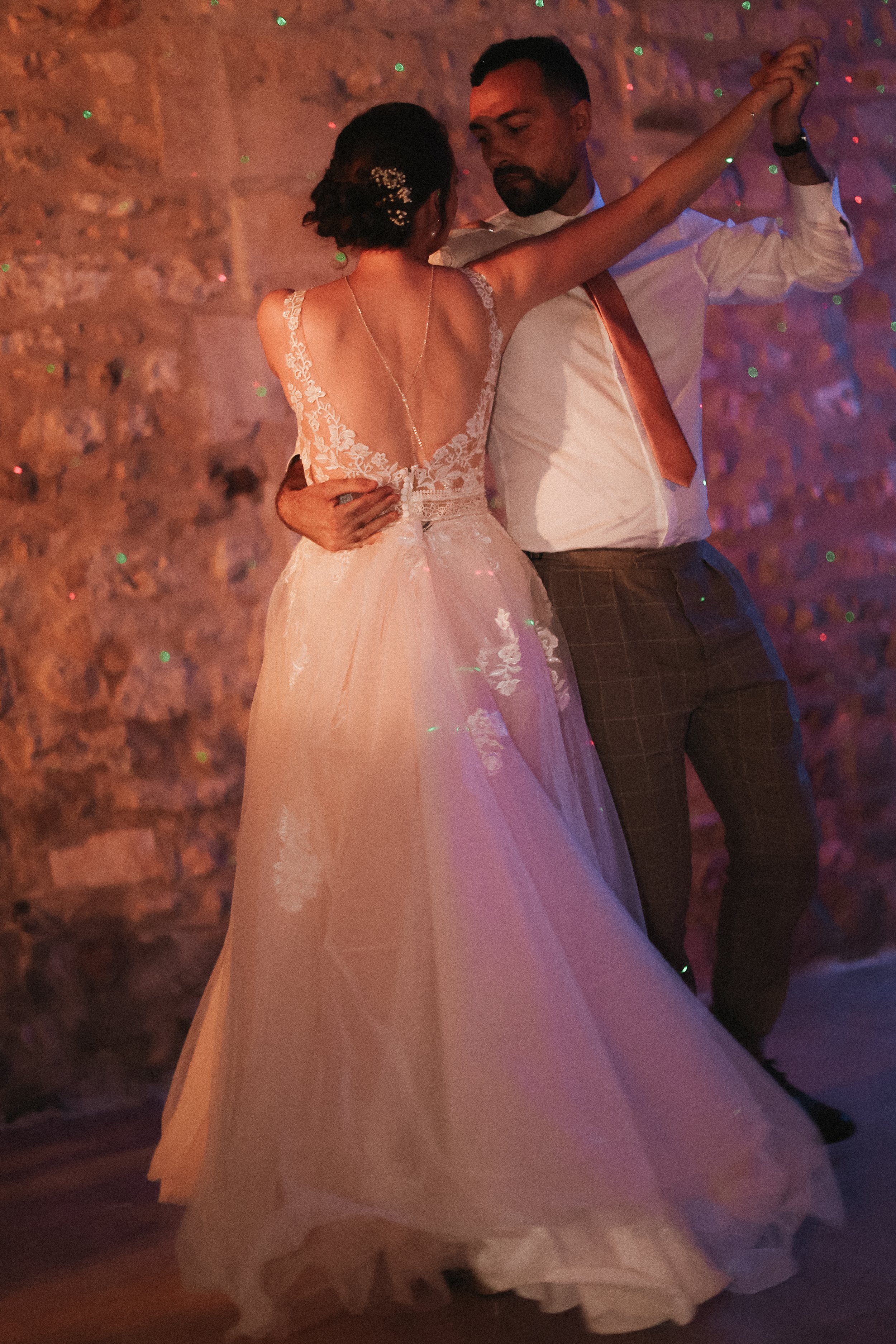 Un couple danse lors d'un mariage, la femme porte une robe de mariée en dentelle et un homme en chemise blanche avec une cravate marron, dans une pièce à mur en pierre.