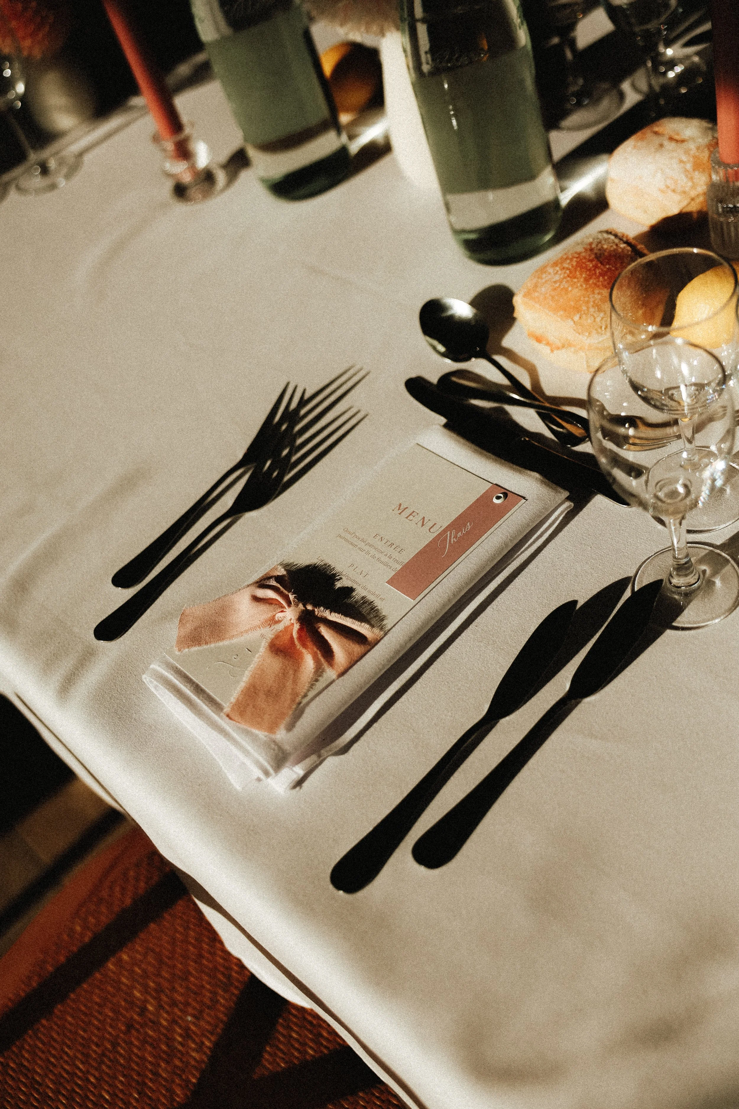 Table dressée avec des couverts noirs, un menu, des verres à vin, du pain et une bouteille de vin dans une ambiance chaleureuse.