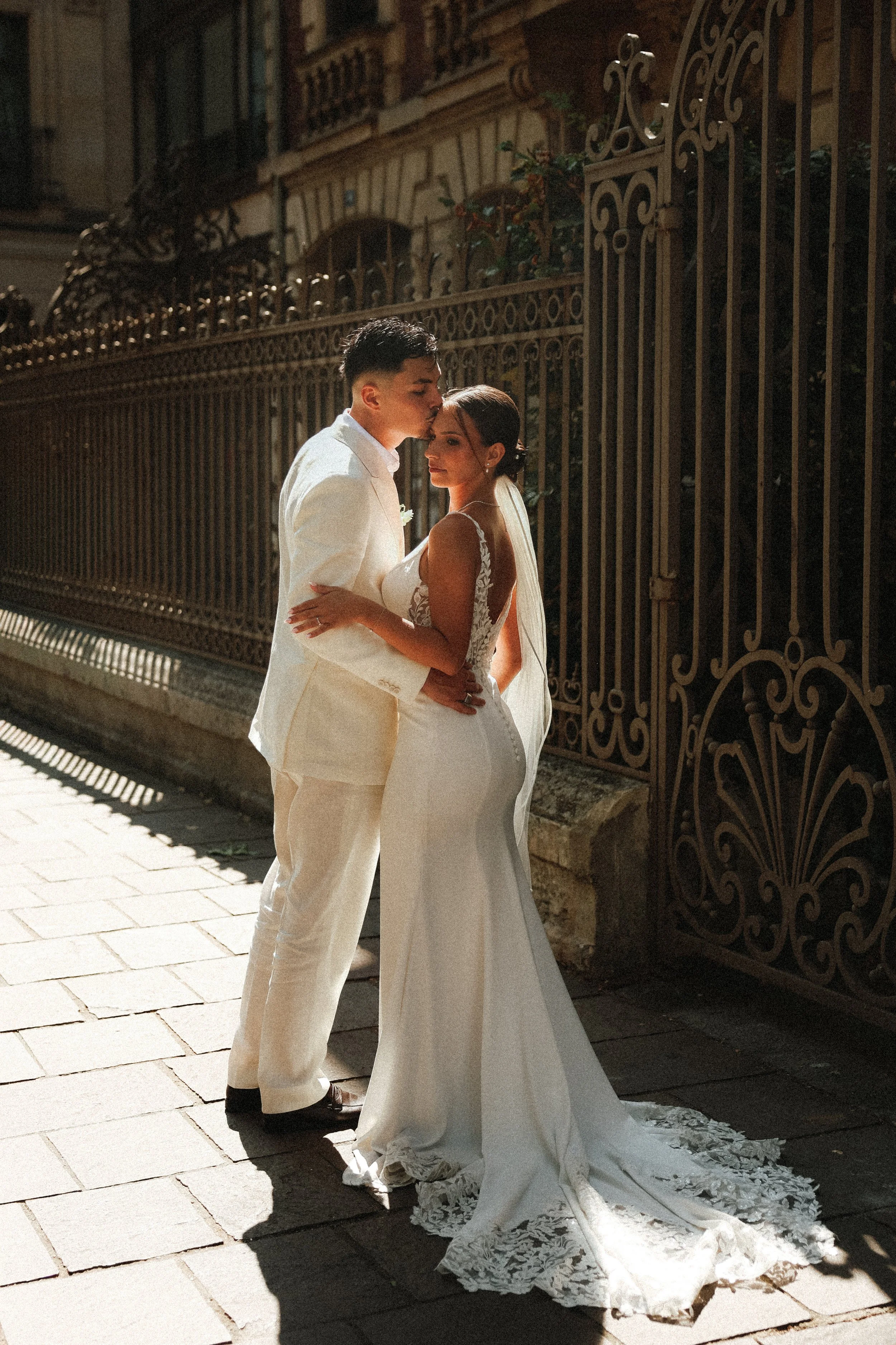 Un couple en mariage sur un trottoir, l'homme en costume blanc et la femme en robe de mariée blanche avec une traîne en dentelle, embrassent devant une clôture en fer forgé, avec des bâtiments anciens en arrière-plan.