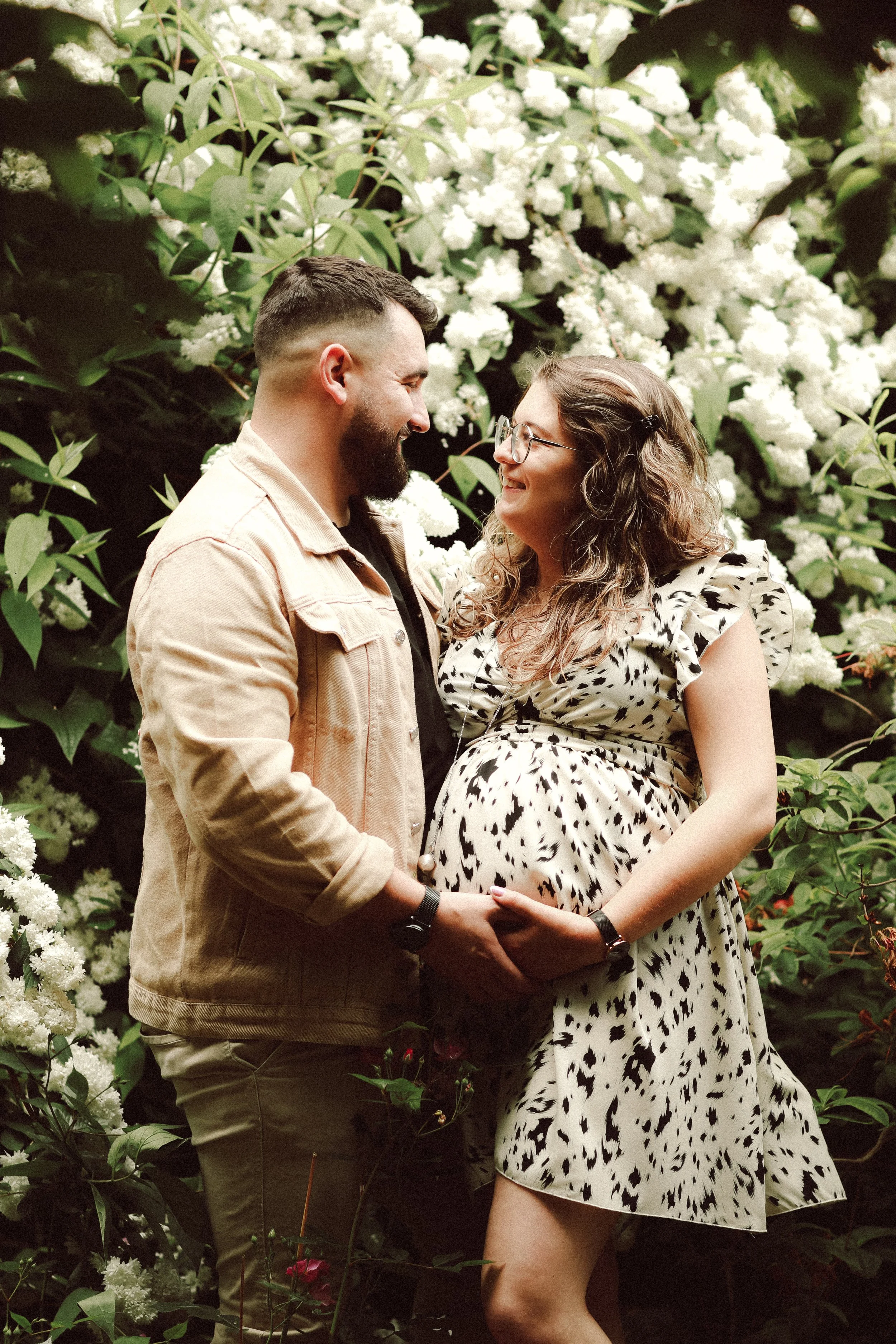 Un couple souriant se tenant par la main devant une haie de buissons blancs en fleurs. La femme est enceinte, portant une robe à imprimé animal et porte des lunettes. L'homme porte une veste beige.
