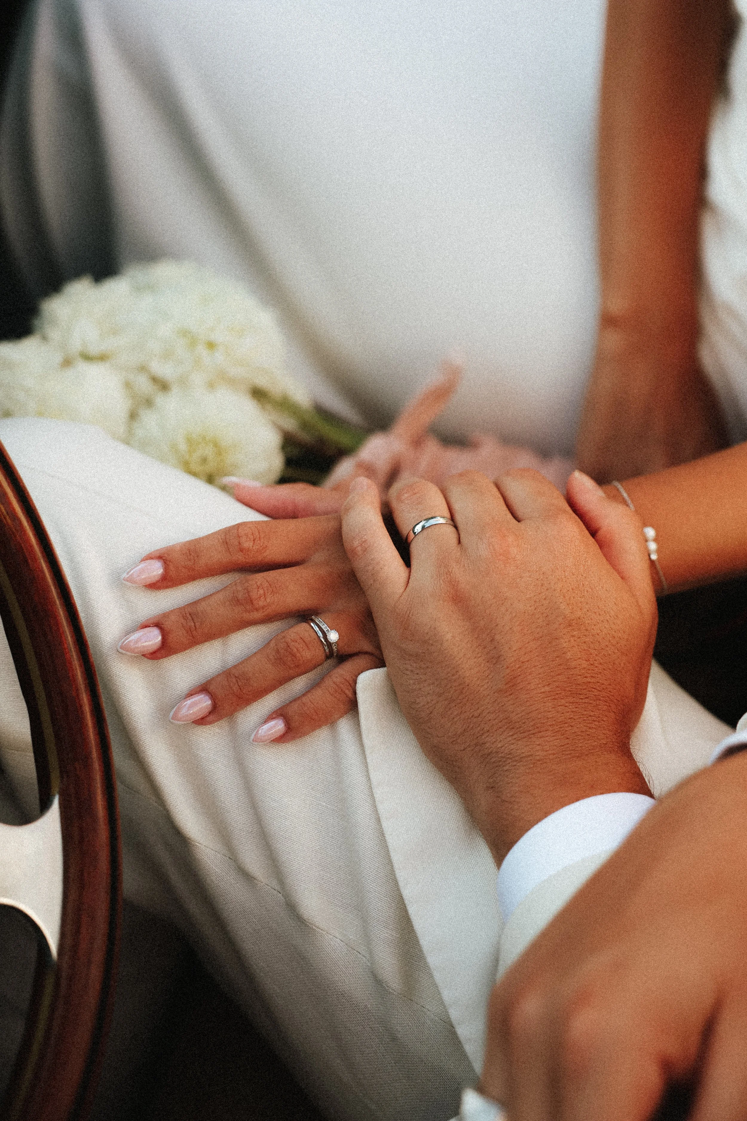 Jeux de mains entre deux personnes portant des alliances, avec un bouquet de fleurs blanches en arrière-plan.