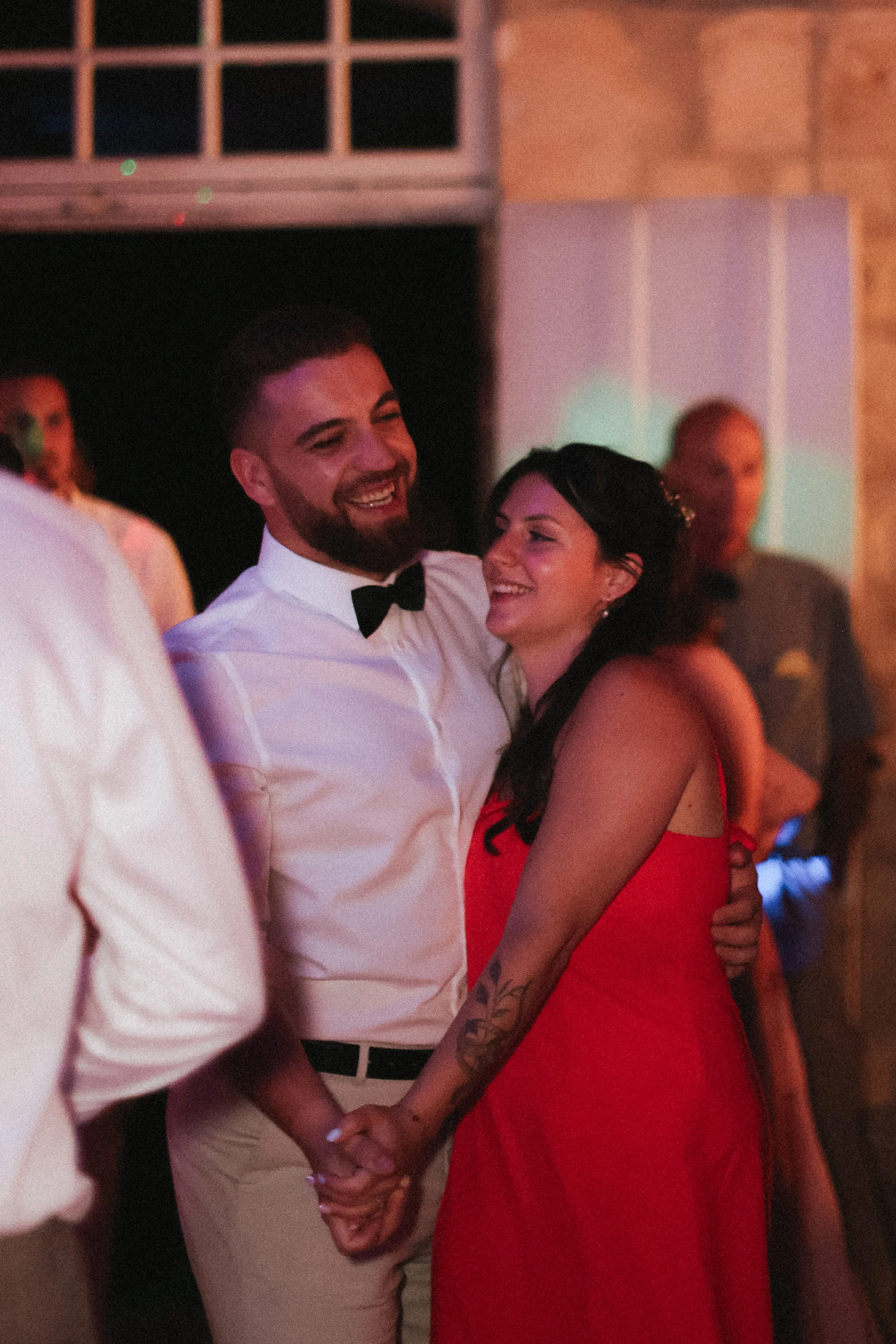 Un couple souriant, tenant la main, lors d'une fête ou d'un bal. La femme porte une robe rouge et des accessoires, l'homme porte une chemise blanche avec nœud papillon. Il y a d'autres personnes en arrière-plan dans une ambiance chaleureuse et festiv