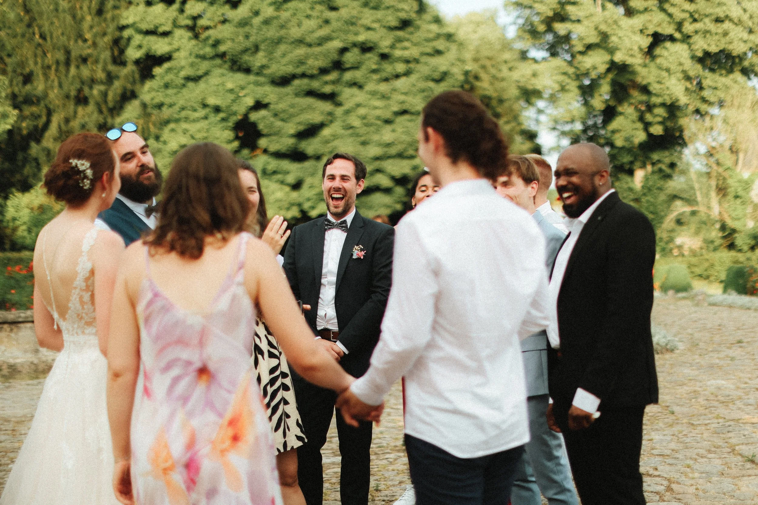 Un groupe de personnes en costume et en robe de mariage se tient par la main en cercle dans un parc, souriant et riant, lors d'une célébration de mariage en extérieur.