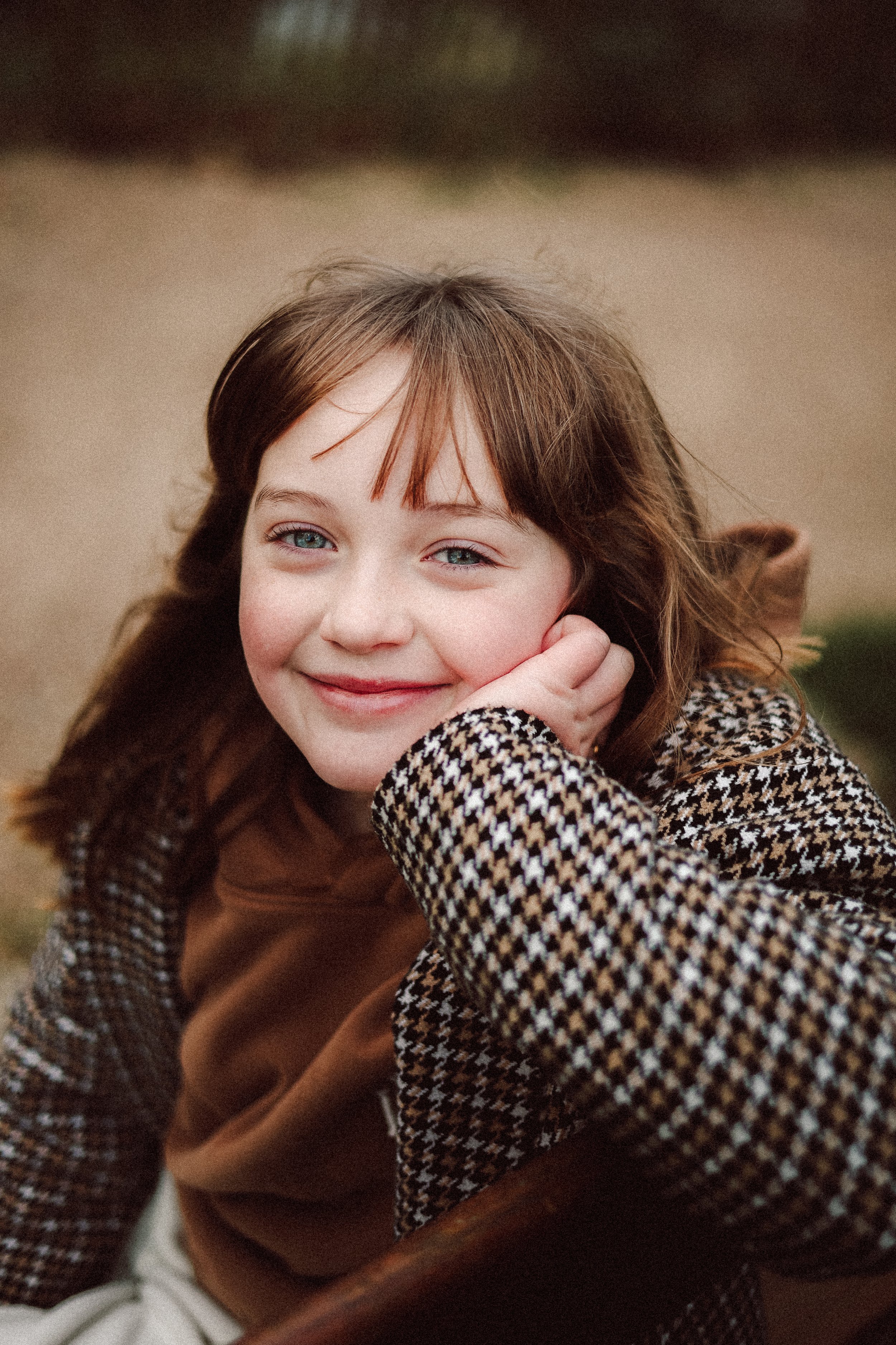 Un garçon souriant avec cheveux longs et bruns, portant un manteau à motif pied-de-poule et une écharpe marron, posé avec la main à la tête, dans un décor extérieur flou.