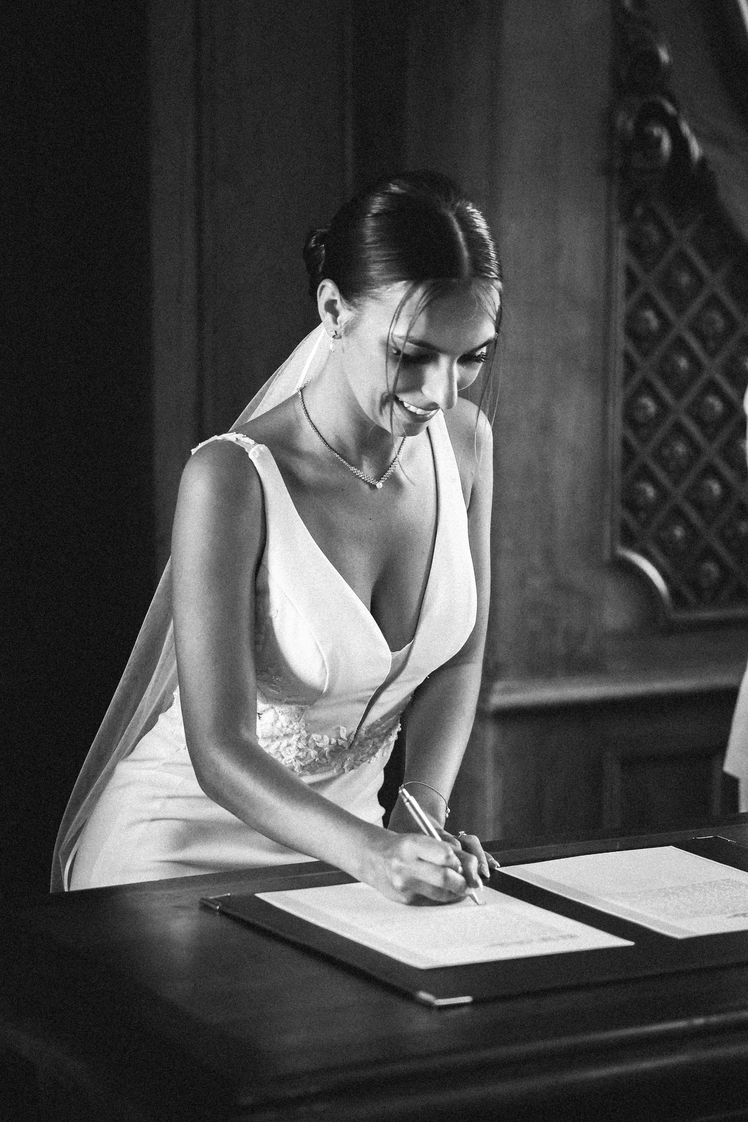 Une femme en robe de mariée signe un document sur un bureau, en noir et blanc.
