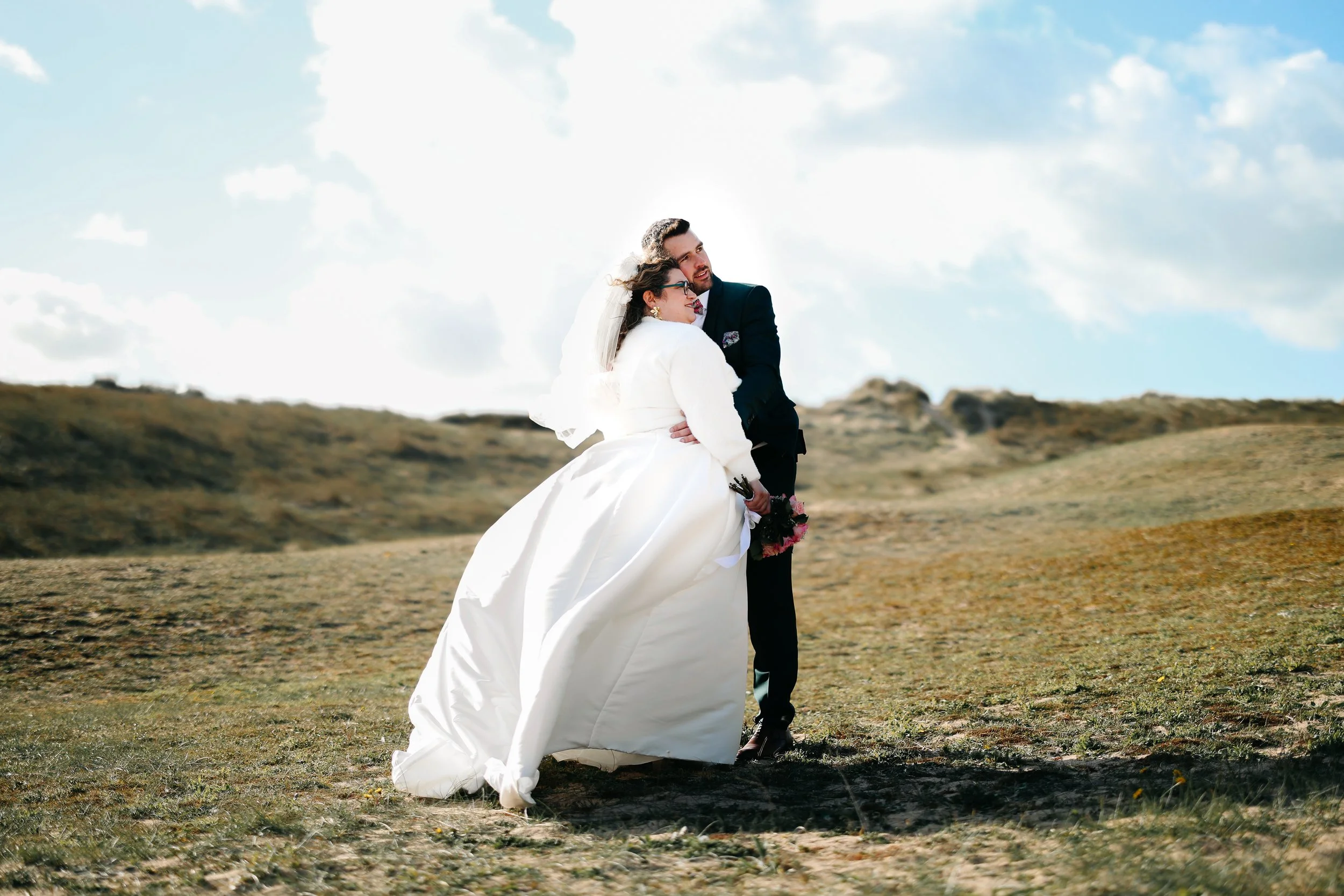 Un couple en habits de mariage, la femme en robe blanche et le homme en costume sombre, se tient dans un paysage naturel avec des collines et un ciel bleu avec des nuages.
