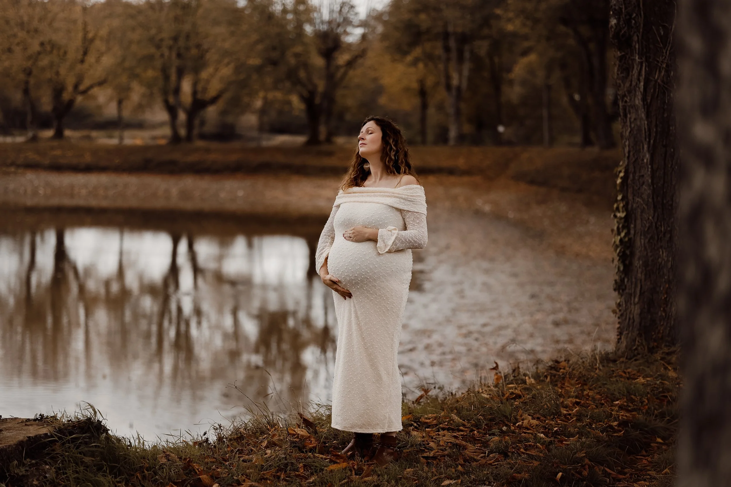 Une femme enceinte en robe blanche debout près d'un lac, dans un paysage automnal avec des arbres aux feuilles jaunes et brunes.