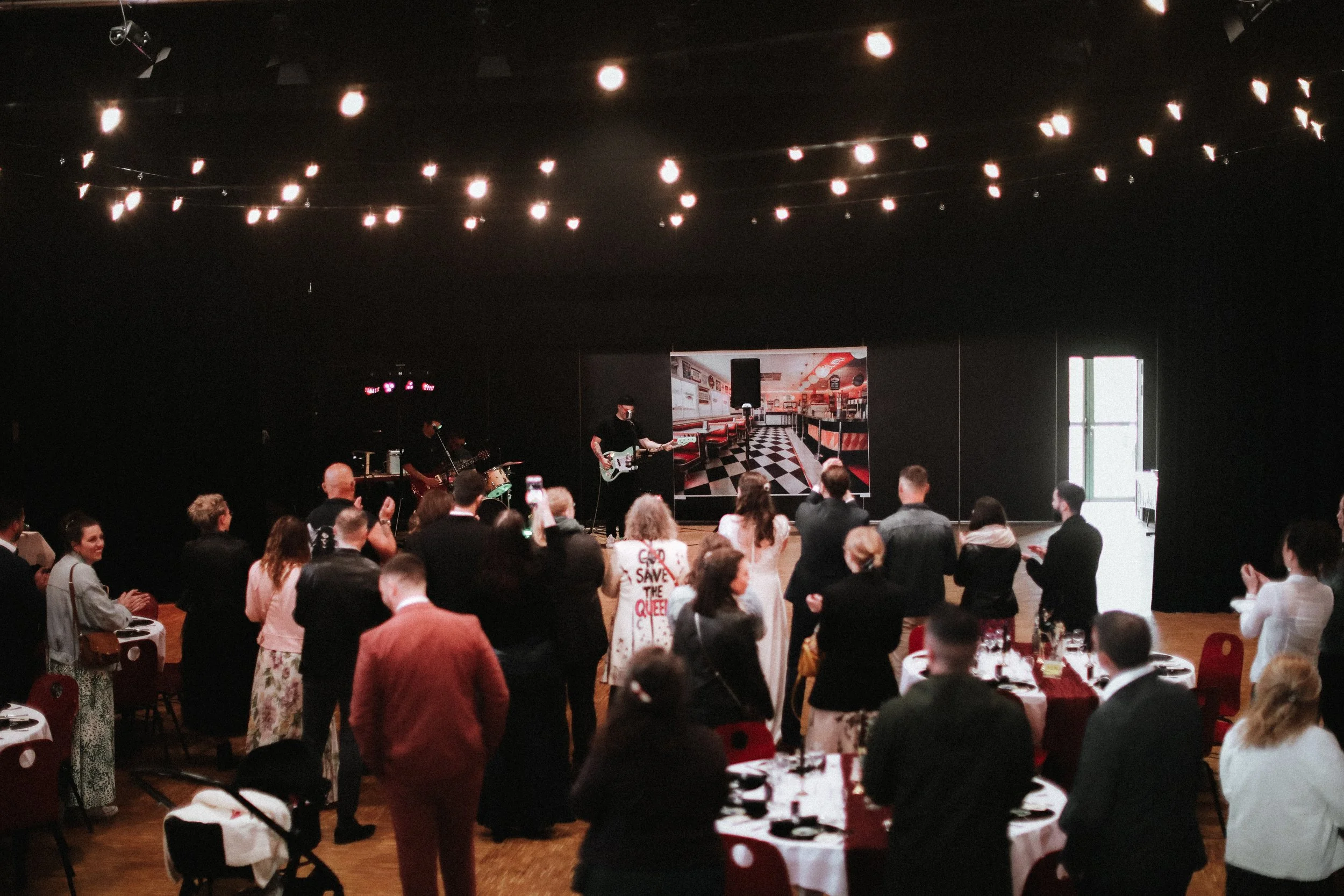 Groupe de personnes assistant à une performance musicale dans une salle sombre avec une scène, des tables et une porte lumineuse, décoration minimaliste