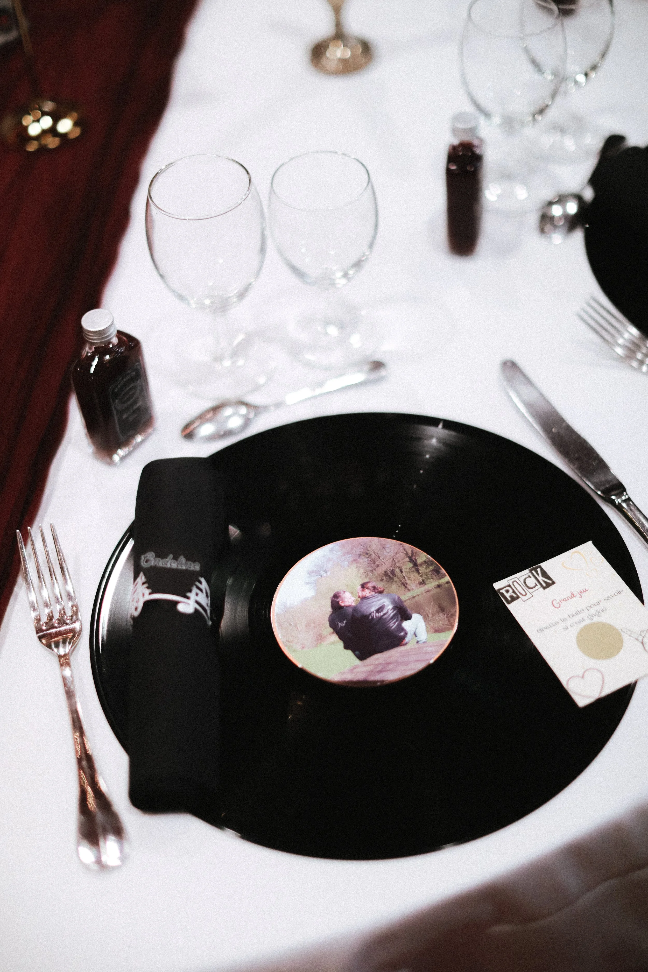 Une table de banquet avec une nappe blanche, un disque en vinyle noir comme centre de table, une photo de deux personnes assises près d'un lac, des verres à vin, des bouteilles, un couteau, une fourchette, et une carte de fête. 