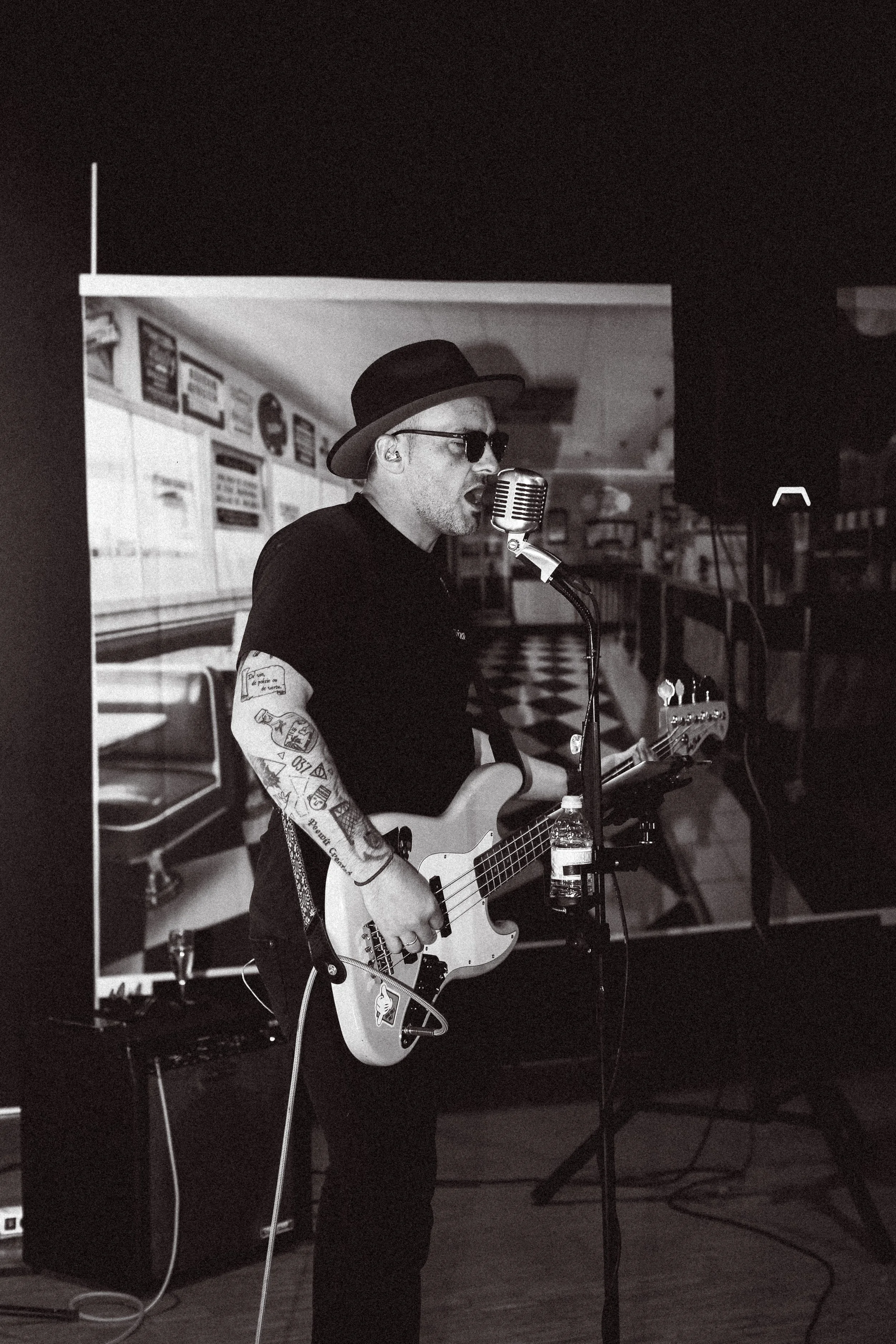 Homme jouant de la guitare et chantant avec un micro, portant un chapeau et des lunettes, dans un lieu de musique ou de performance, en noir et blanc.