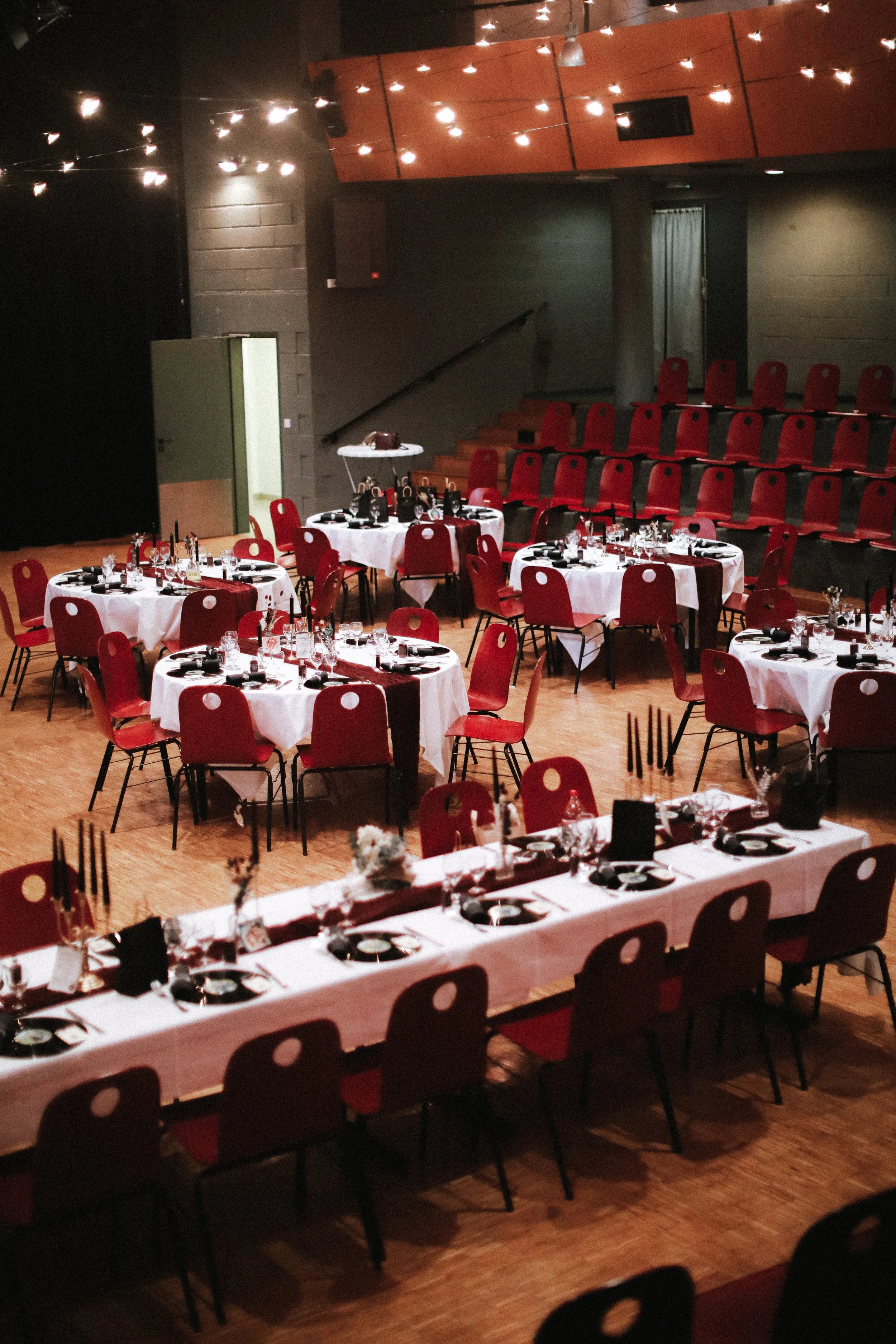 Salle de réception avec tables rondes et une longue table, décorée pour un événement, avec chaises rouges, nappes blanches, bougies et verres, ambiance chaleureuse et élégante.