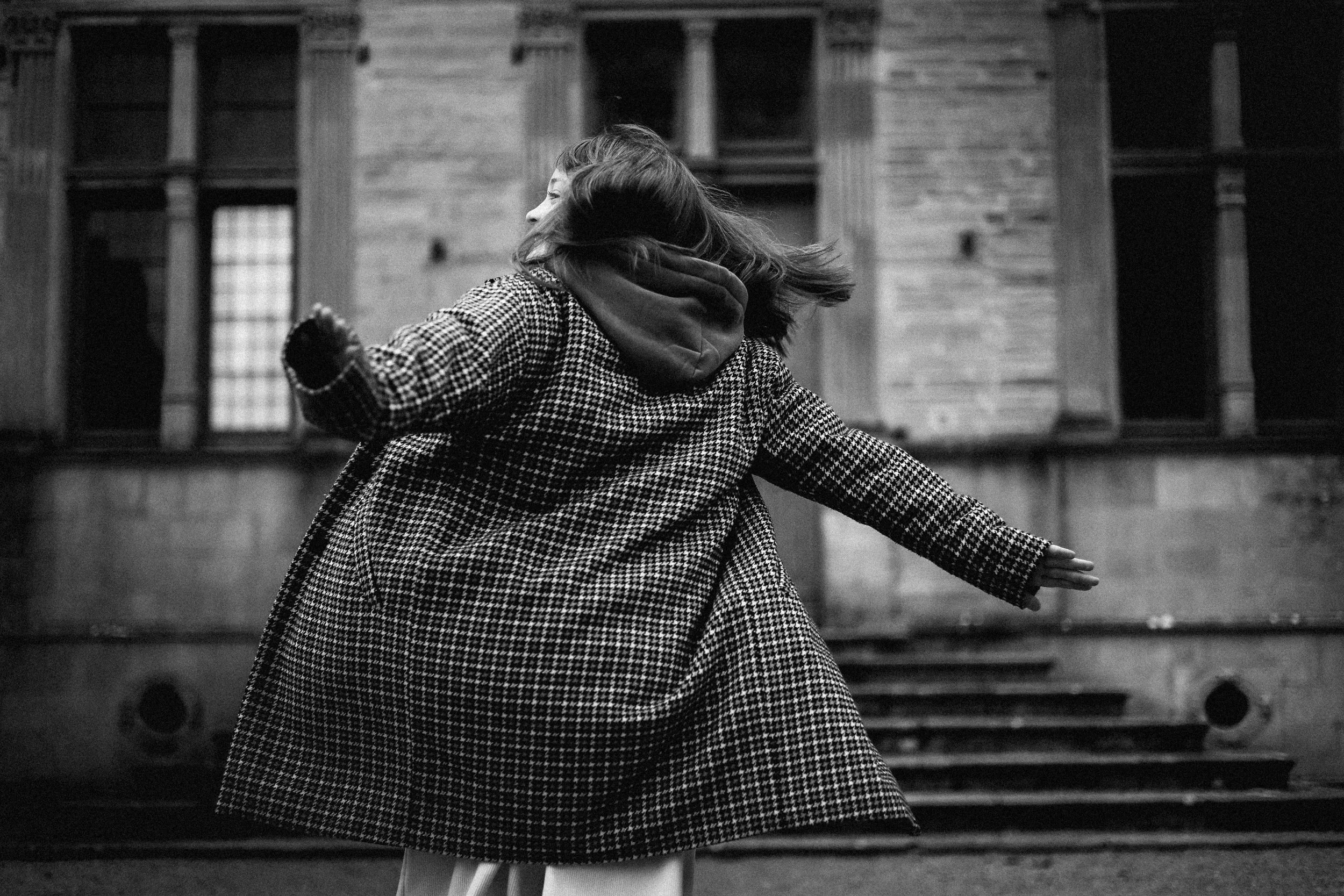 Femme en manteau à carreaux et capuche, tournant et levant un bras dans une rue urbaine, en noir et blanc.
