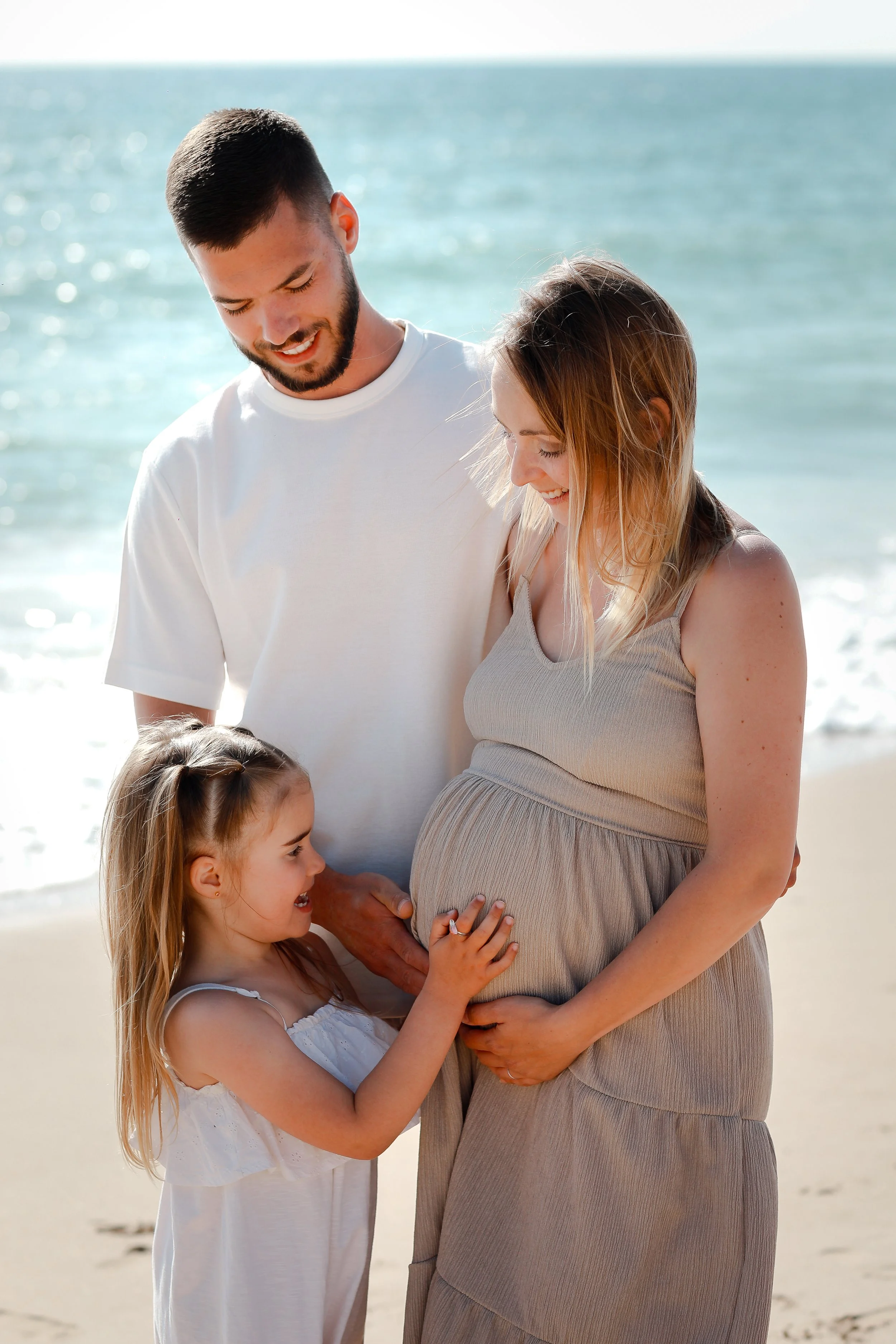 Un homme, une femme enceinte et deux jeunes filles à la plage, avec la mer en arrière-plan, partageant un moment affectueux.