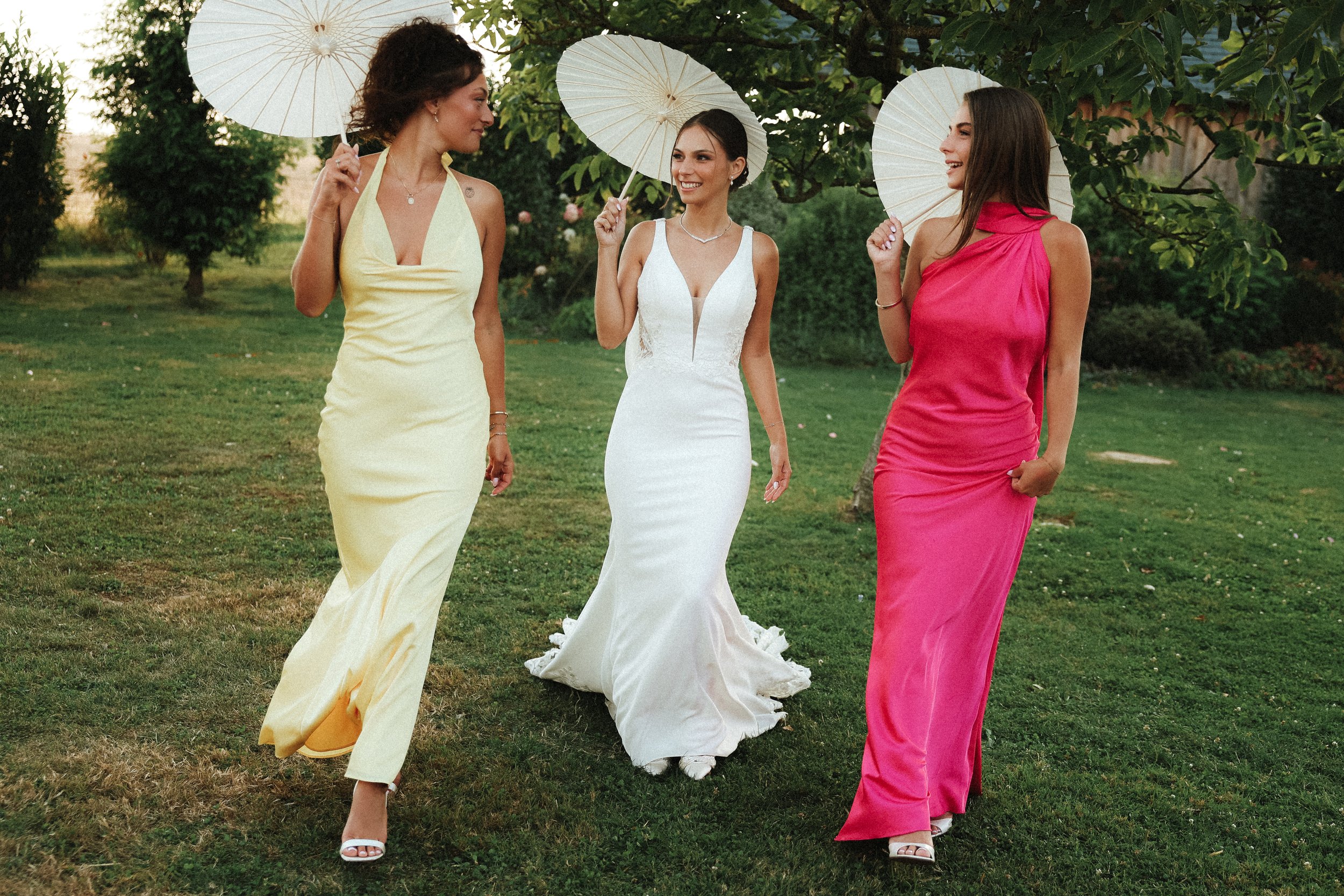 Trois femmes élégantes portent des robes longues dans un jardin, tenant des parapluies blancs, en train de discuter et de sourire.
