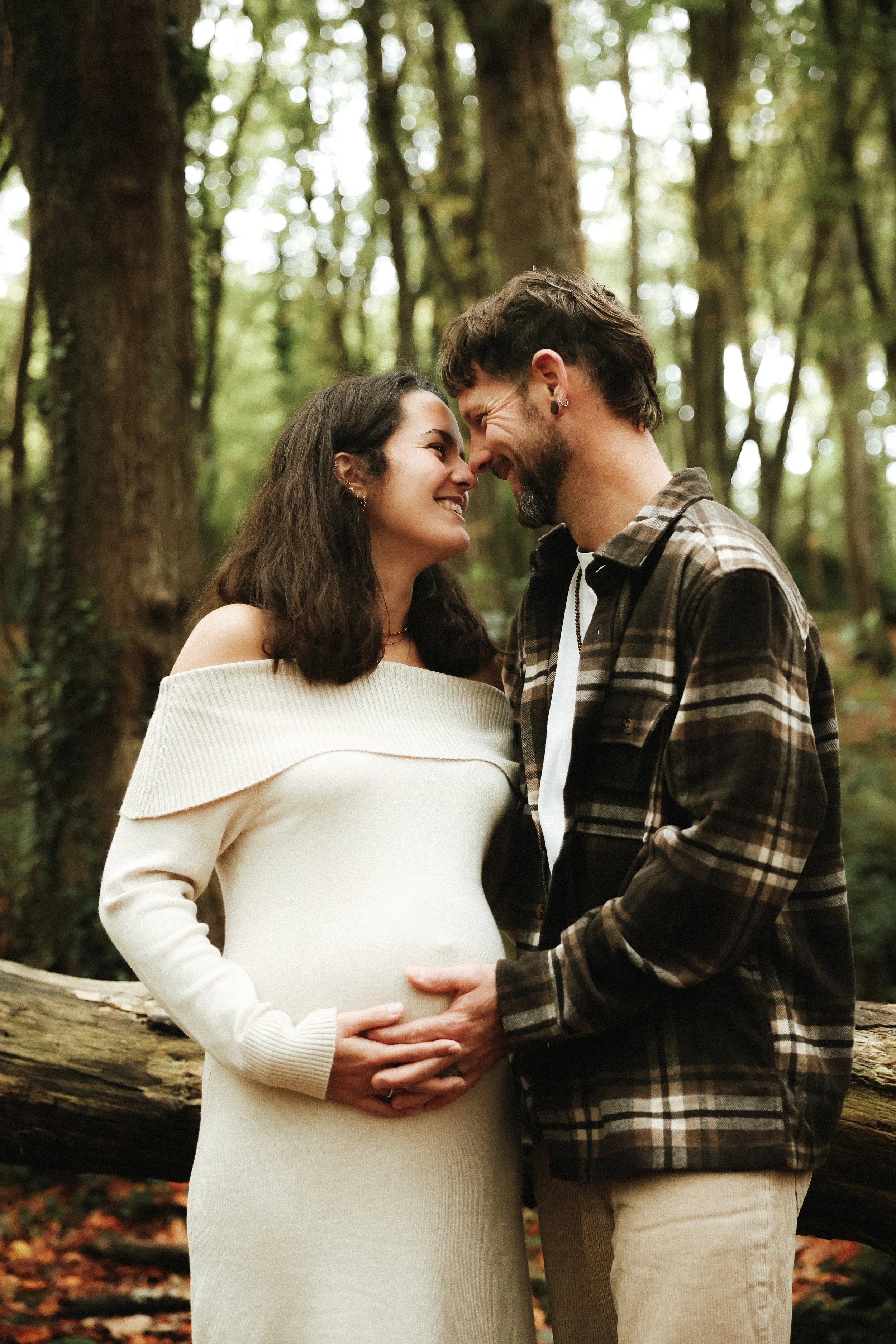 Un couple en forêt, la femme enceinte et le mari se tiennent contre un arbre, se touchent le front et s'embrassent tendrement.