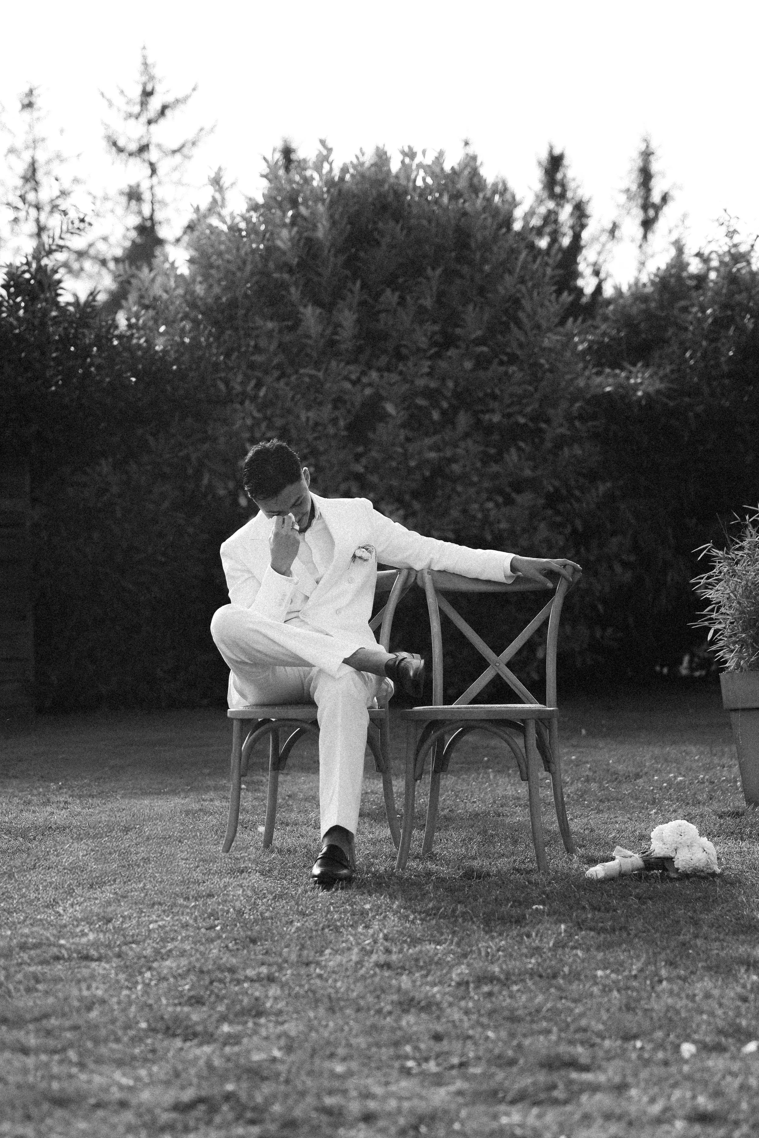 Un homme habillé en blanc assis sur une chaise en extérieur, l'air pensif, avec des arbres en arrière-plan, en noir et blanc.