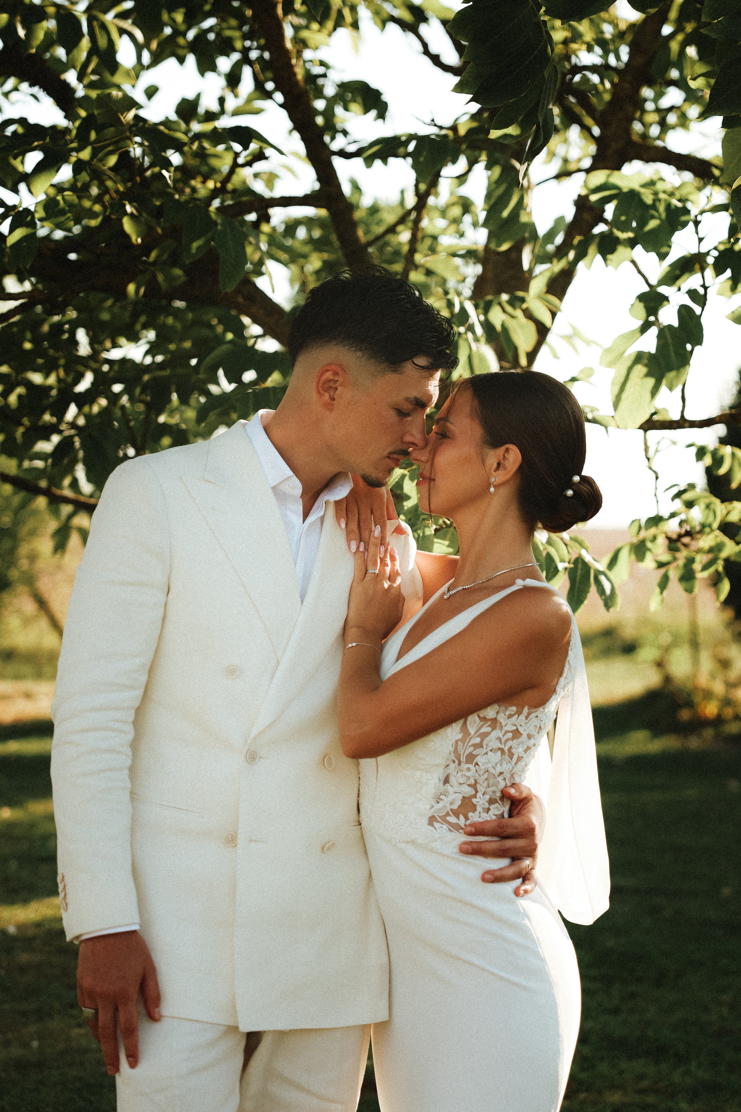 Un couple de mariés en robe blanche et costume blanc, s'embrassant tendrement à l'extérieur sous un arbre, lors de leur mariage.