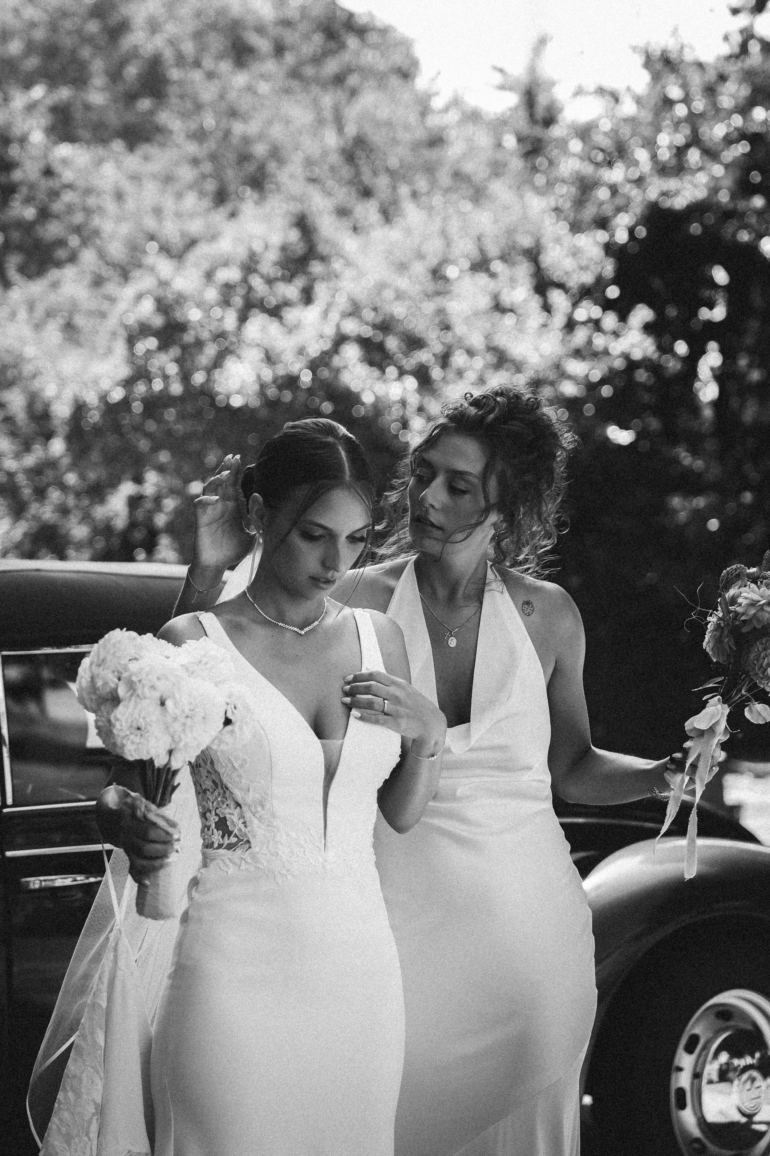 Deux femmes en robes de mariage, l'une tenant un bouquet de fleurs, la scène semble être une préparation nuptiale en extérieur.