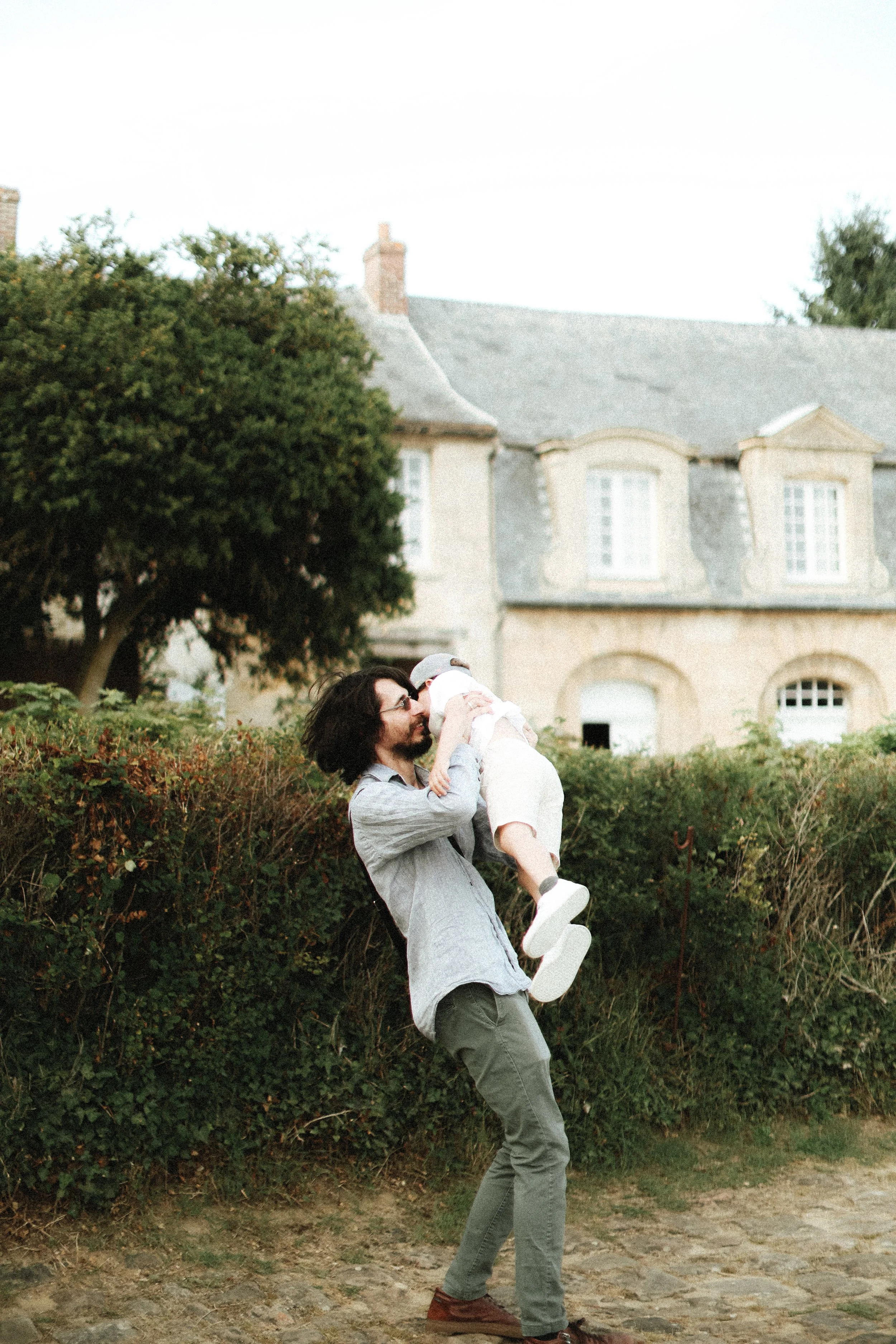 Un homme avec des cheveux longs tient un enfant dans ses bras en plein air devant une maison en pierre avec des fenêtres blanches.