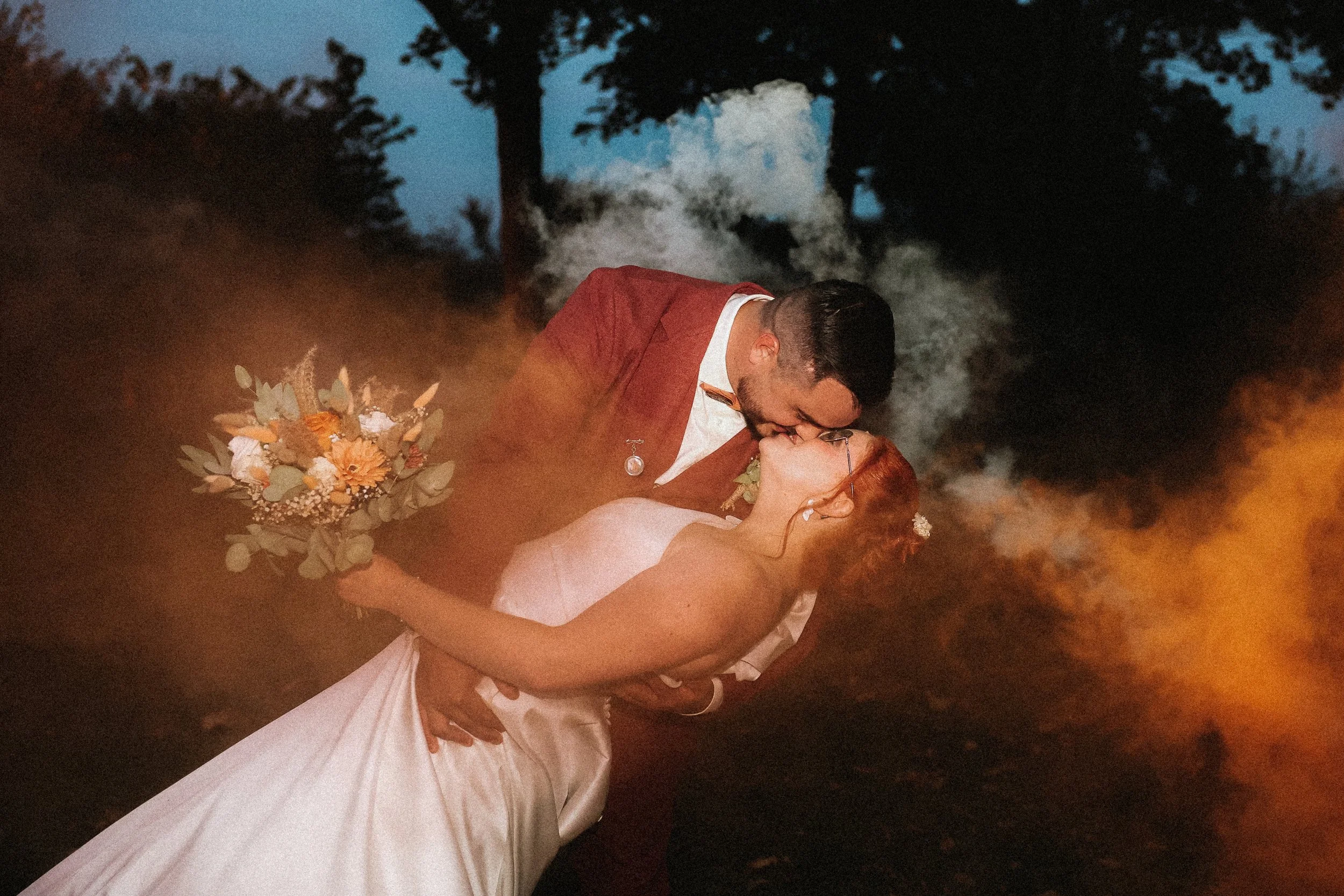 Un couple de mariés dans une scène romantique en extérieur, avec des effets de fumée ou de couleur orange, partageant un baiser passionné. La femme porte une robe blanche et tient un bouquet de fleurs multicolores, tandis que l'homme porte un costume rouge.