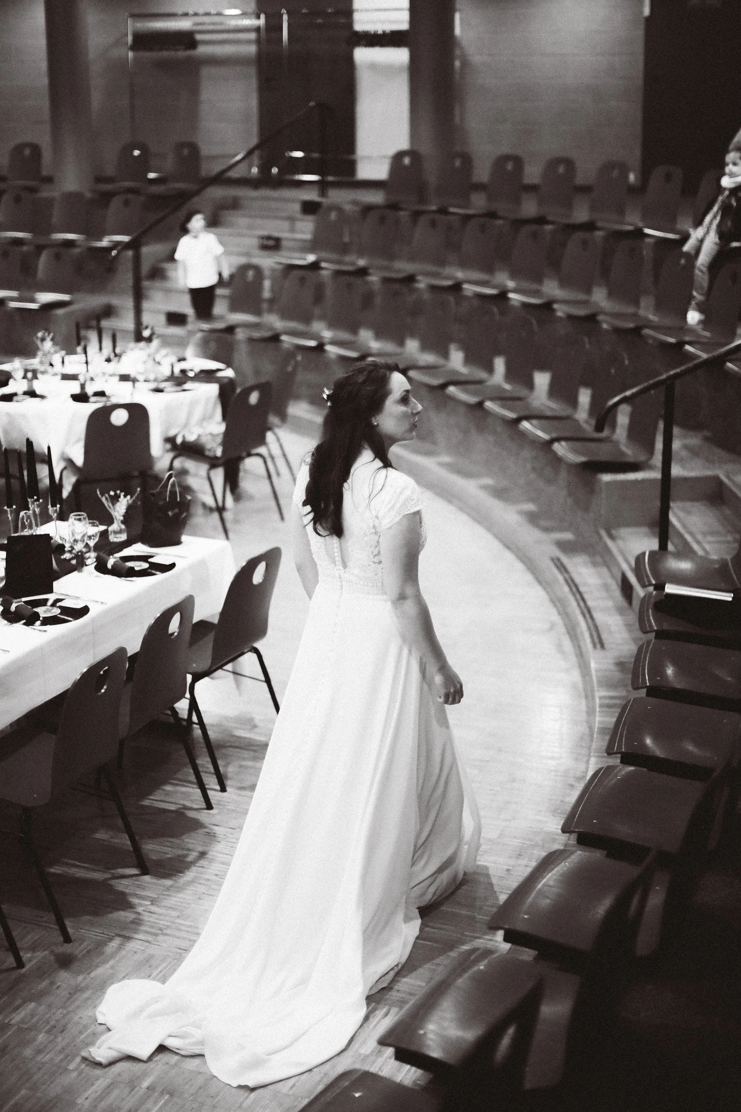 Une femme en robe de mariée dans une salle de banquet vide, accompagnée de deux enfants, l'un en arrière et l'autre en haut des gradins.