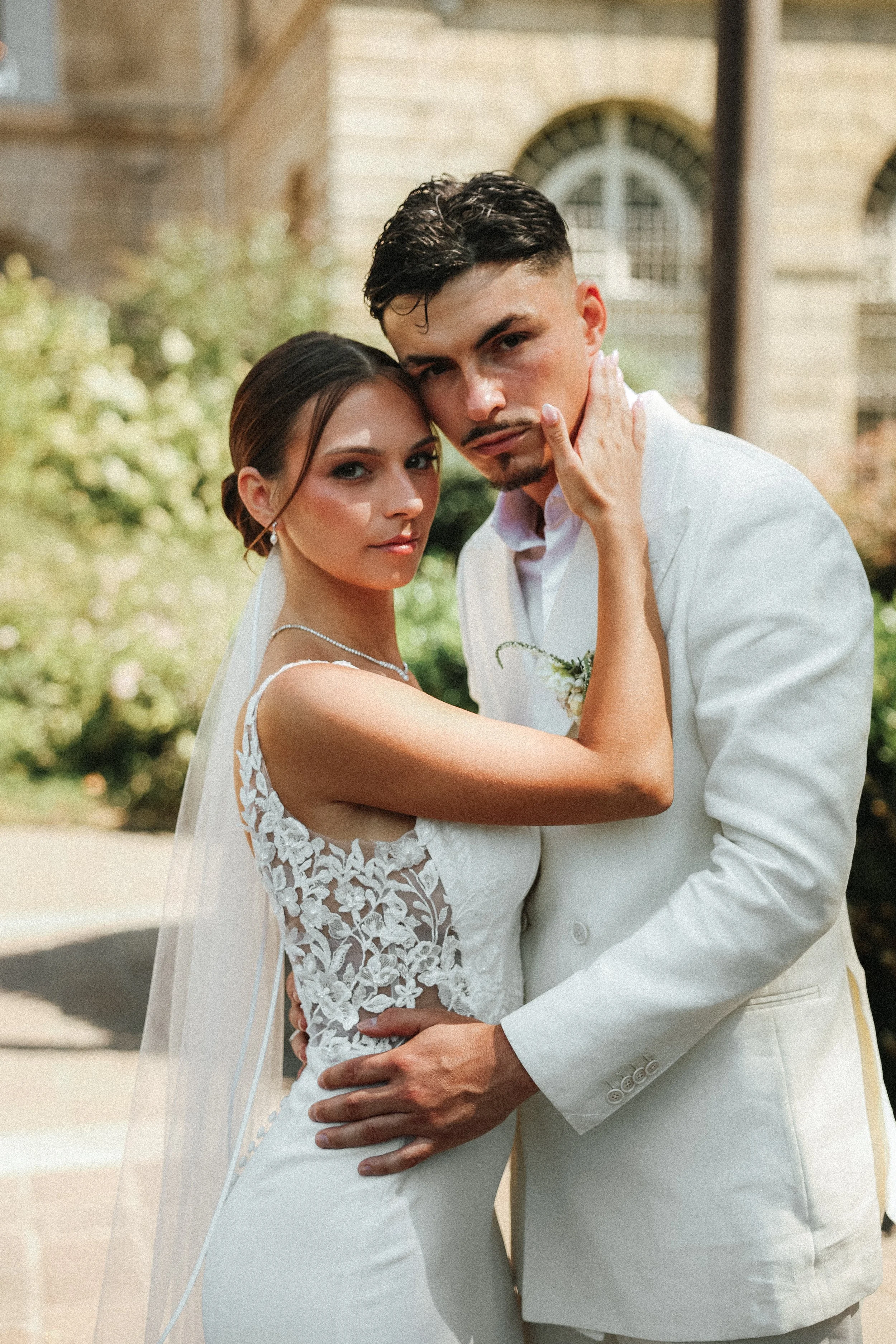 Un couple en mariage pose en extérieur, la femme en robe blanche avec dentelle et le mari en costume blanc, avec un bâtiment ancien en arrière-plan.