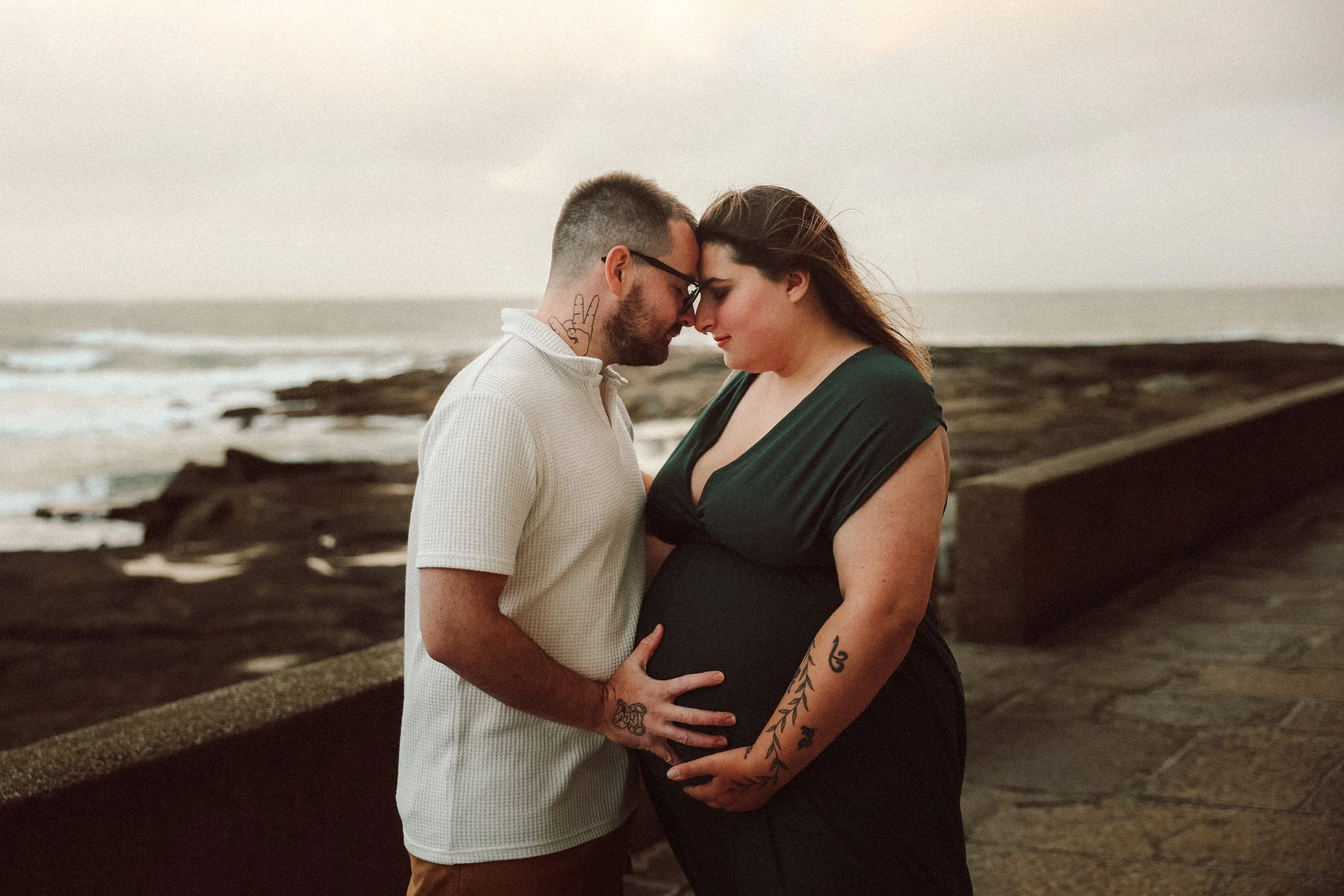 Un couple jeune, un homme et une femme, en train de s'embrasser tendrement, avec la tête appuyée l'un contre l'autre, au bord de la mer au coucher du soleil, avec des tatouages visibles sur leurs bras et leur main de l'homme sur le ventre de la femme